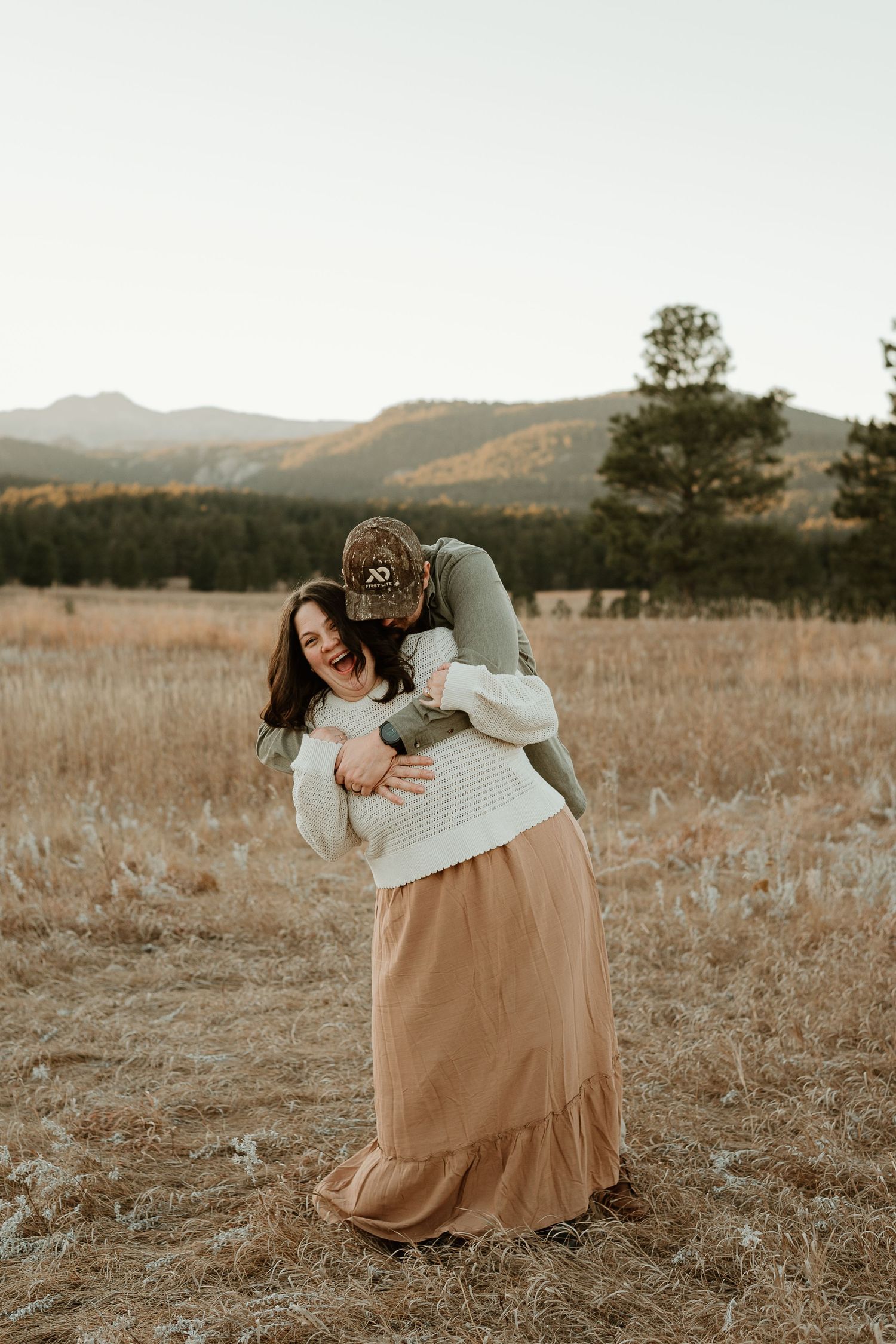 Intimate couple embraces in a golden field with mountains and evergreen trees in the distance at sunset.