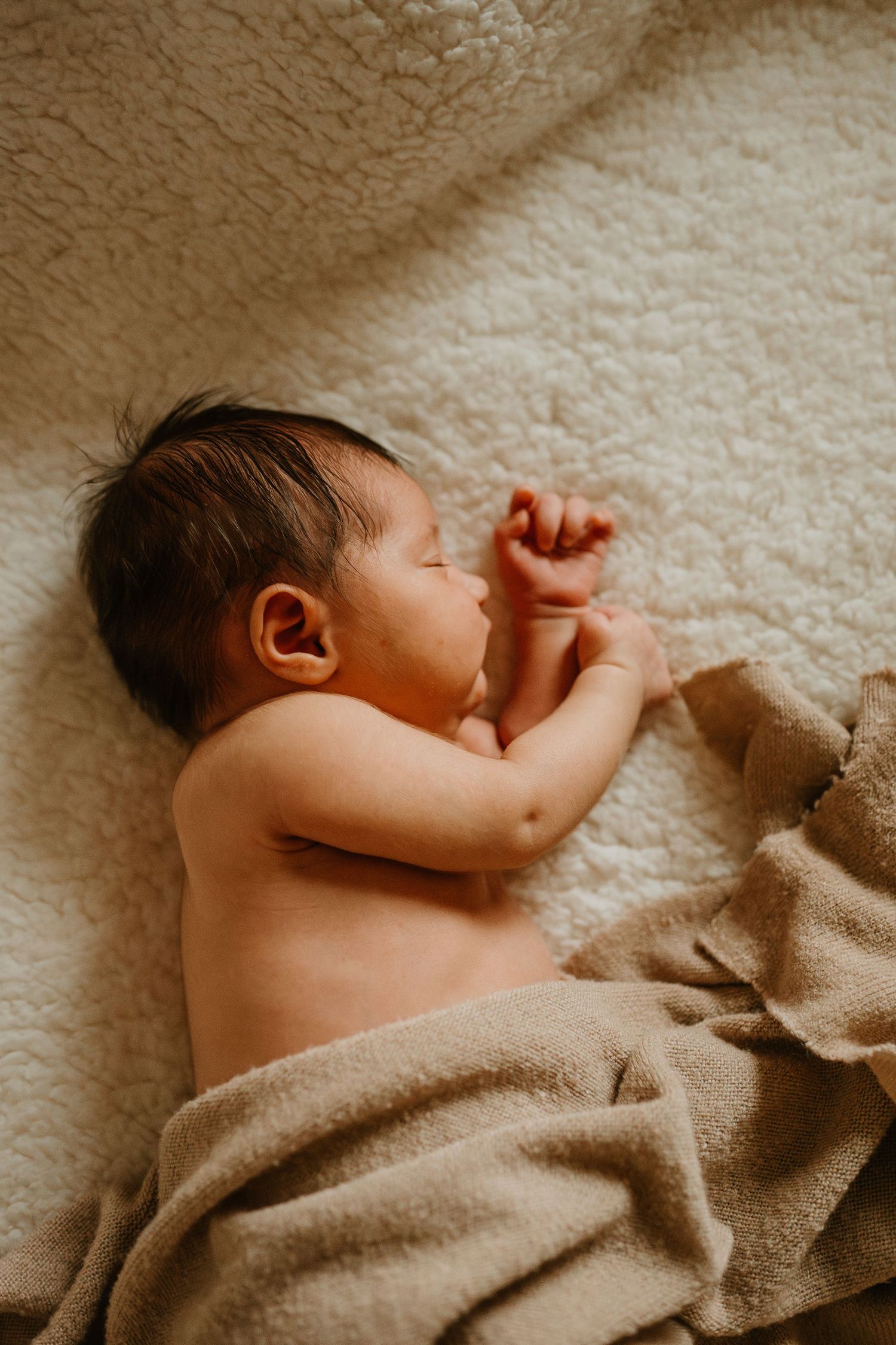 A sleeping newborn baby lies peacefully on a cream-colored blanket in warm lighting.