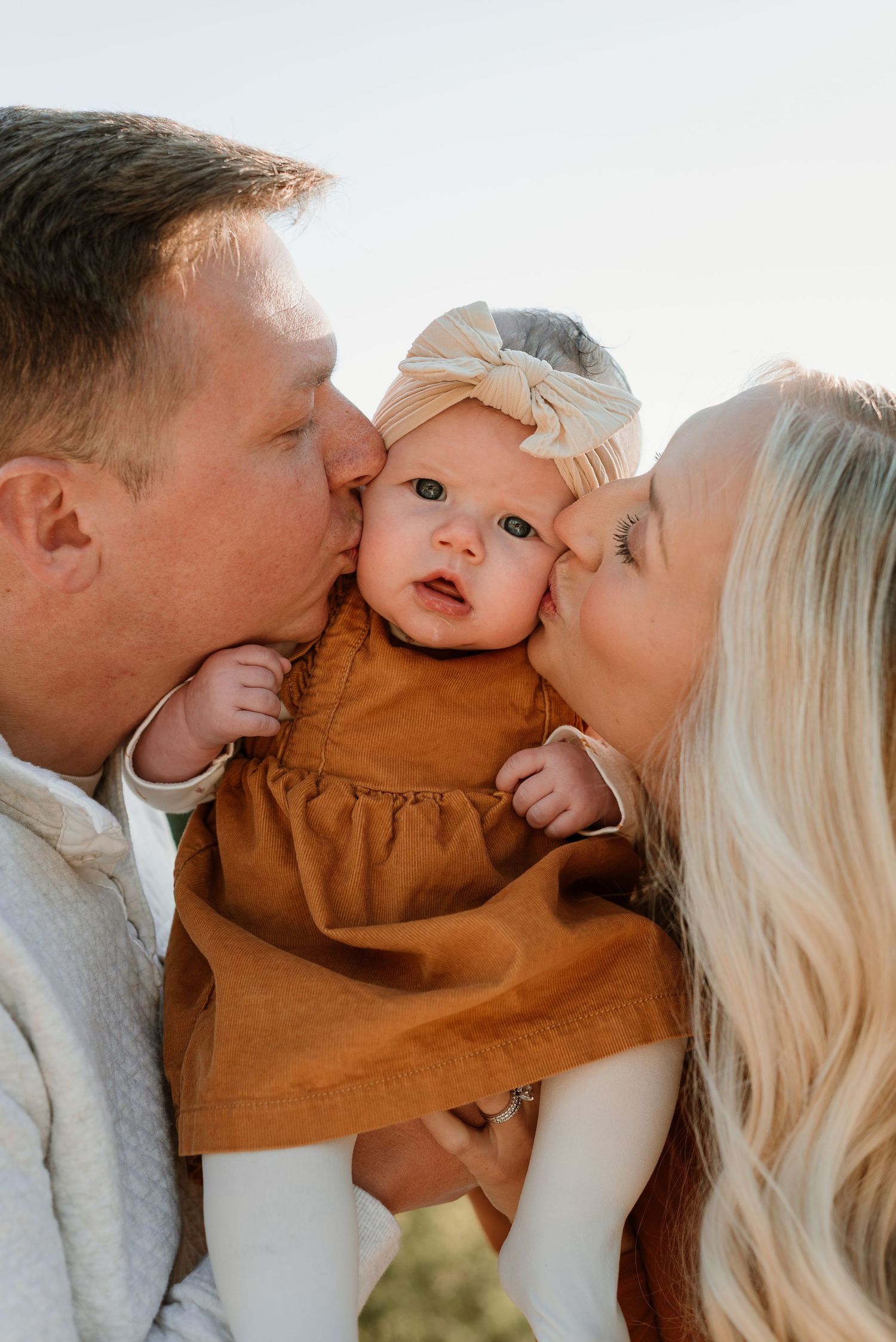 A baby in a brown dress and bow headband gets kisses from both sides in a sweet family moment.