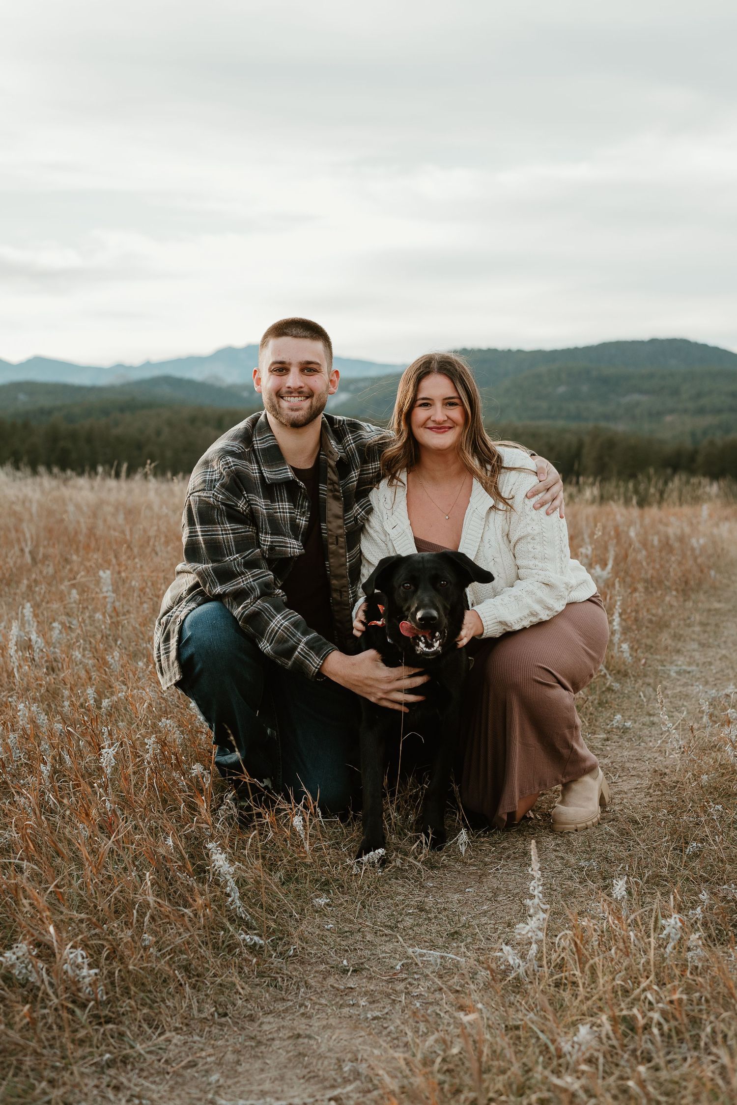 Couple sits in autumn field with their black dog against mountain backdrop in series of intimate outdoor portraits.