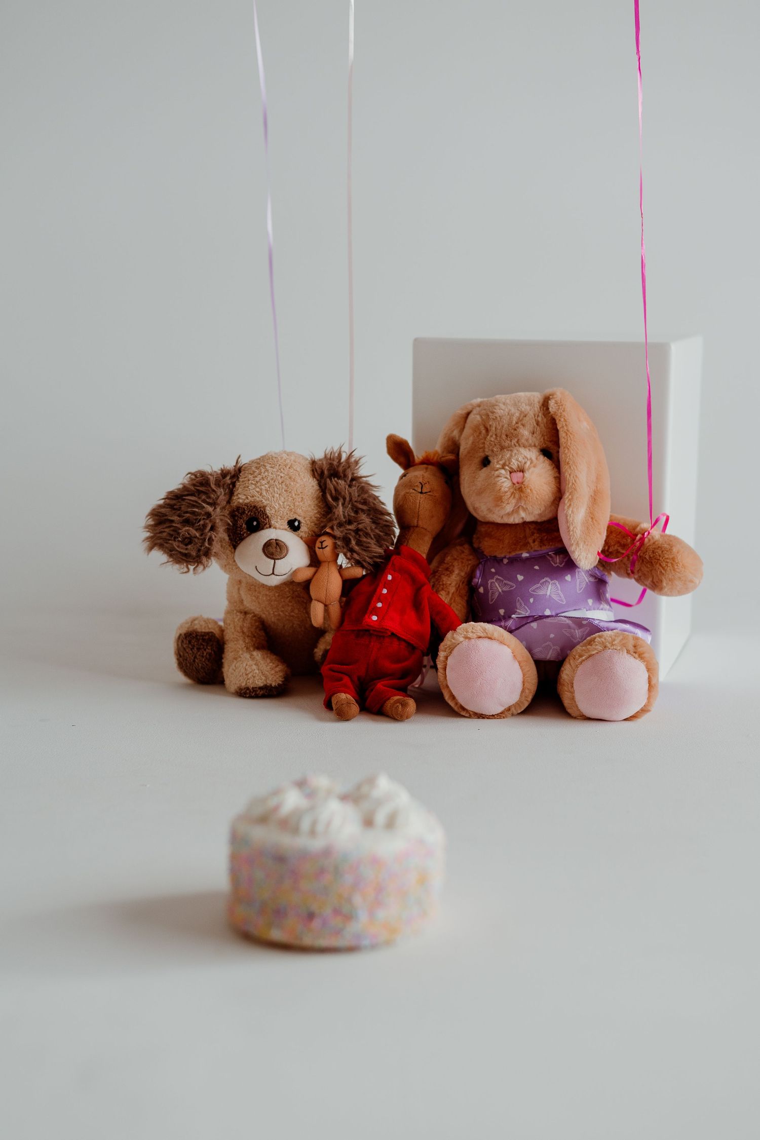 Three plush stuffed animals including a brown dog, pink bunny, and teddy bear sit on a light colored surface.