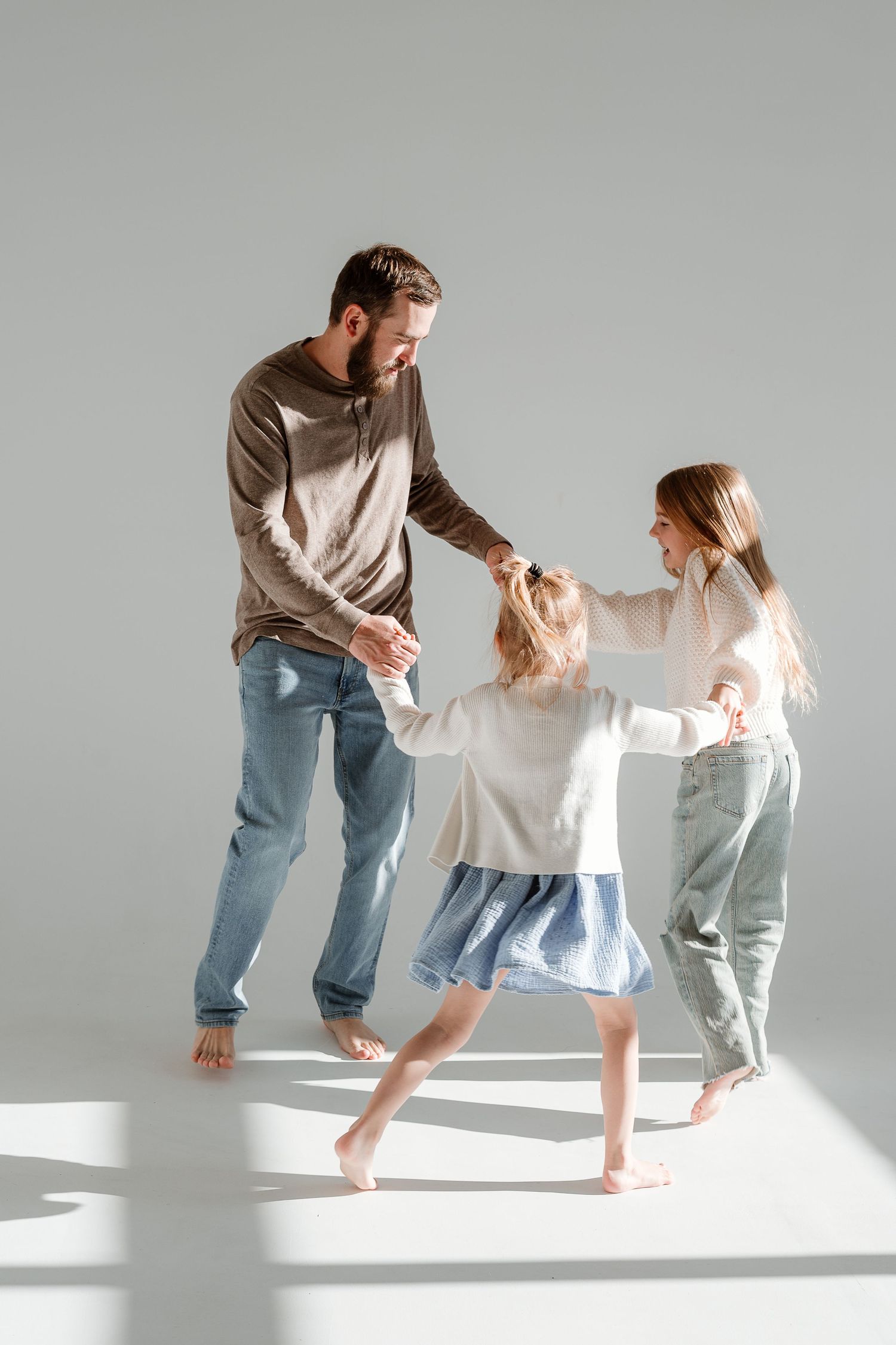 A family dances together barefoot in a bright minimalist setting wearing casual clothing.
