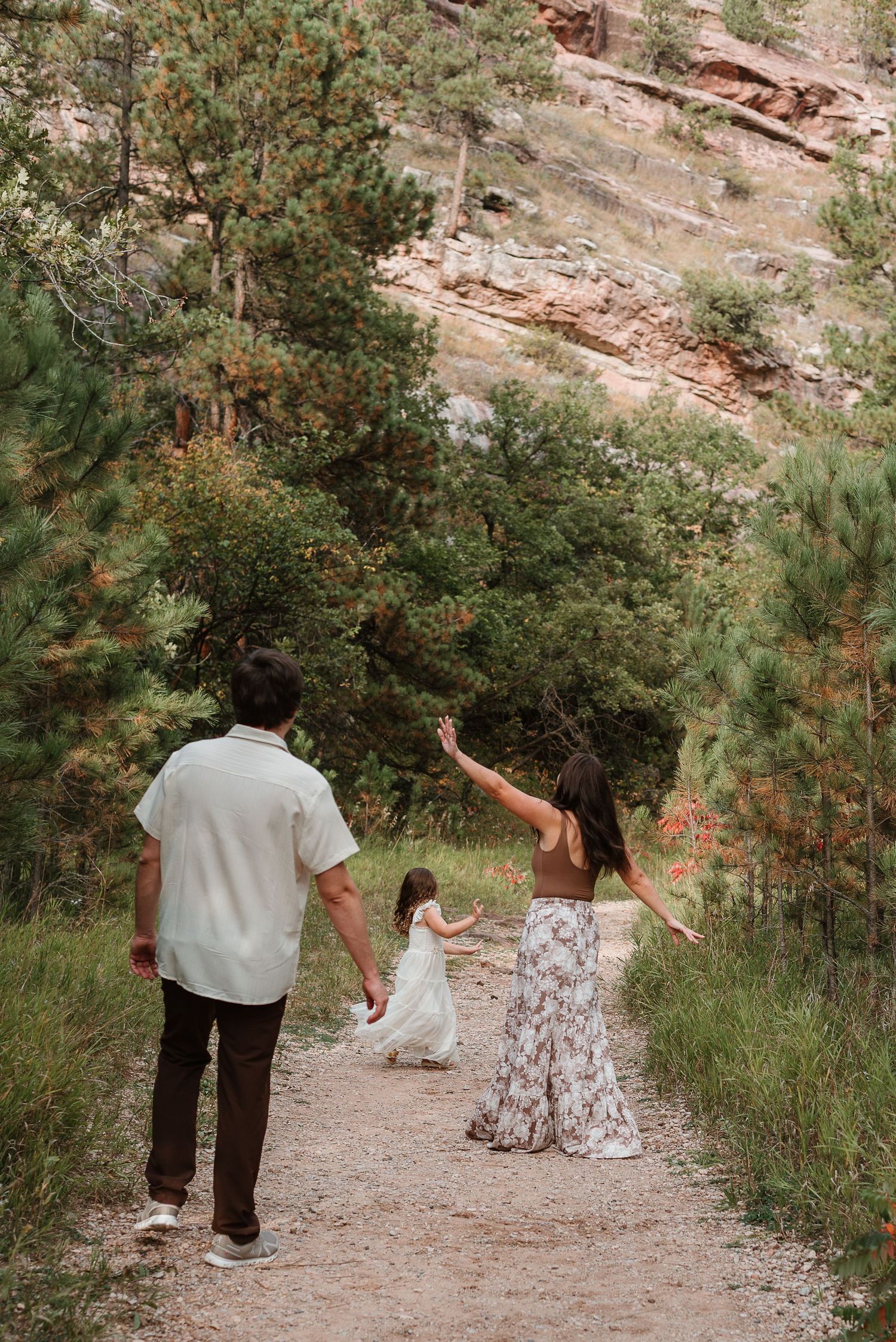 A family walks together on a scenic mountain trail surrounded by pine trees and rocky cliffs.