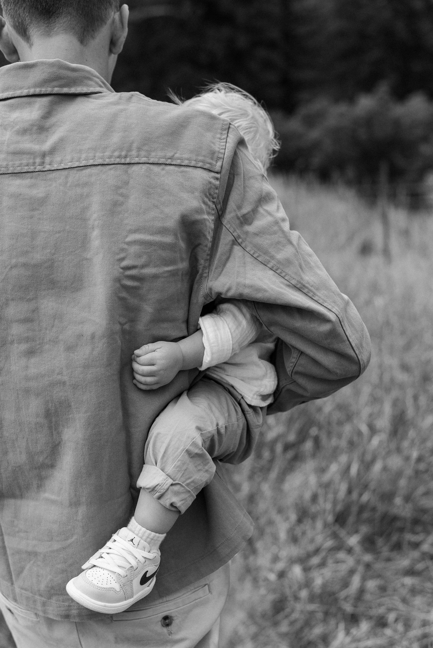 Black and white photo of a parent carrying a small child wearing Nike sneakers in their jacket.