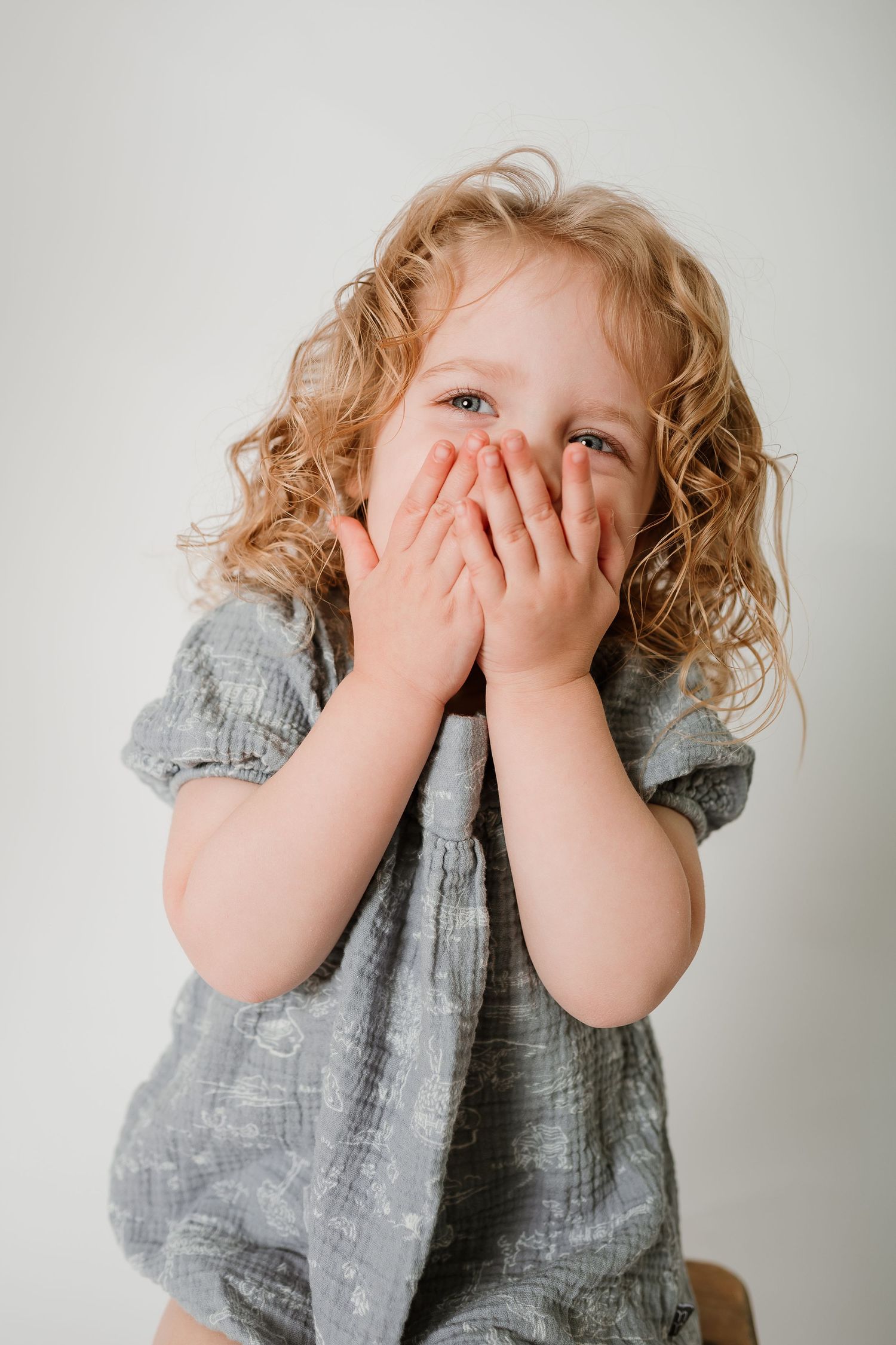 Young child with curly blonde hair wearing a gray dress covers mouth with hands while looking up playfully.