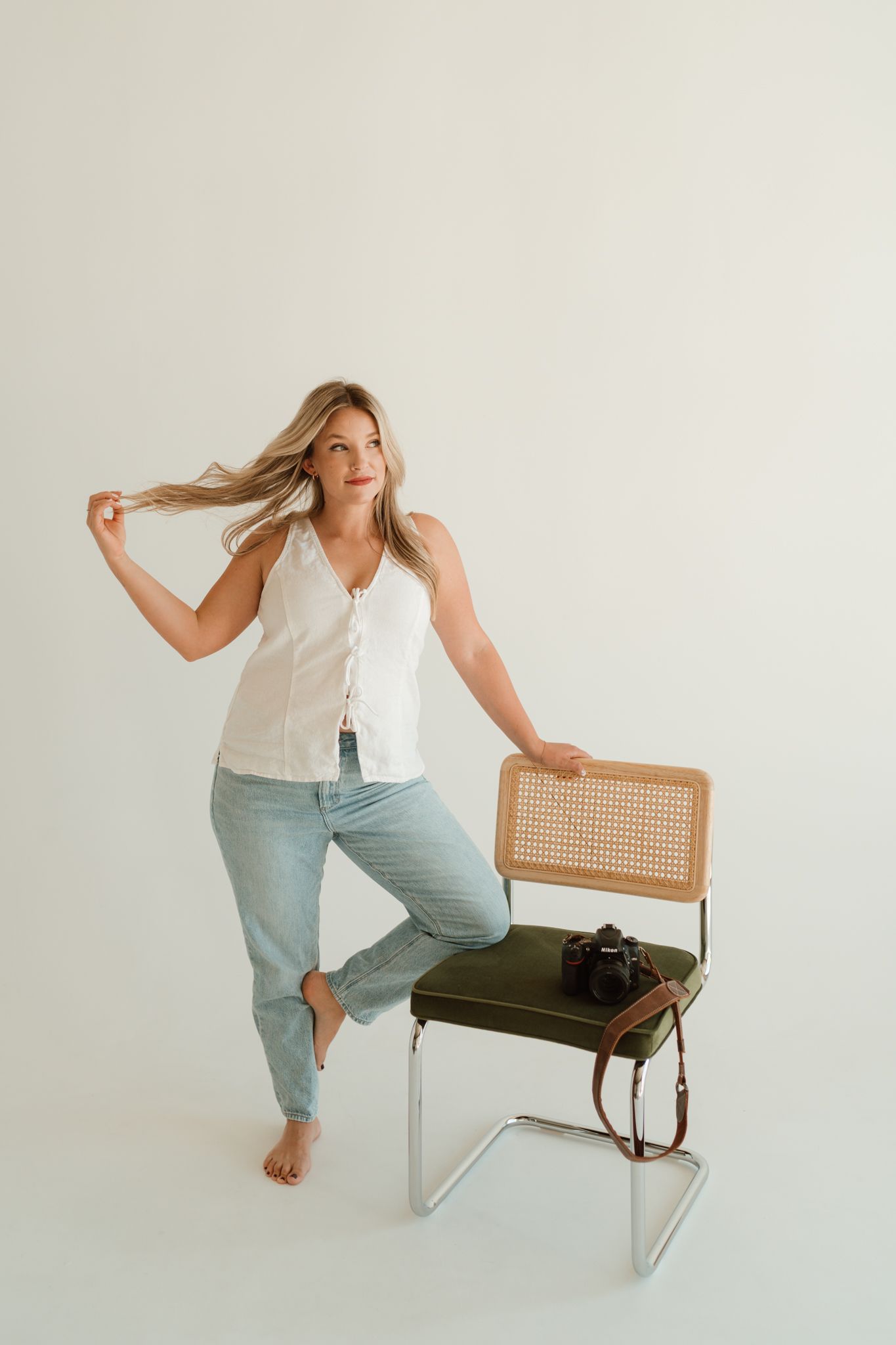 Casual lifestyle portrait with white button-down top, light jeans and vintage cane chair against neutral background.