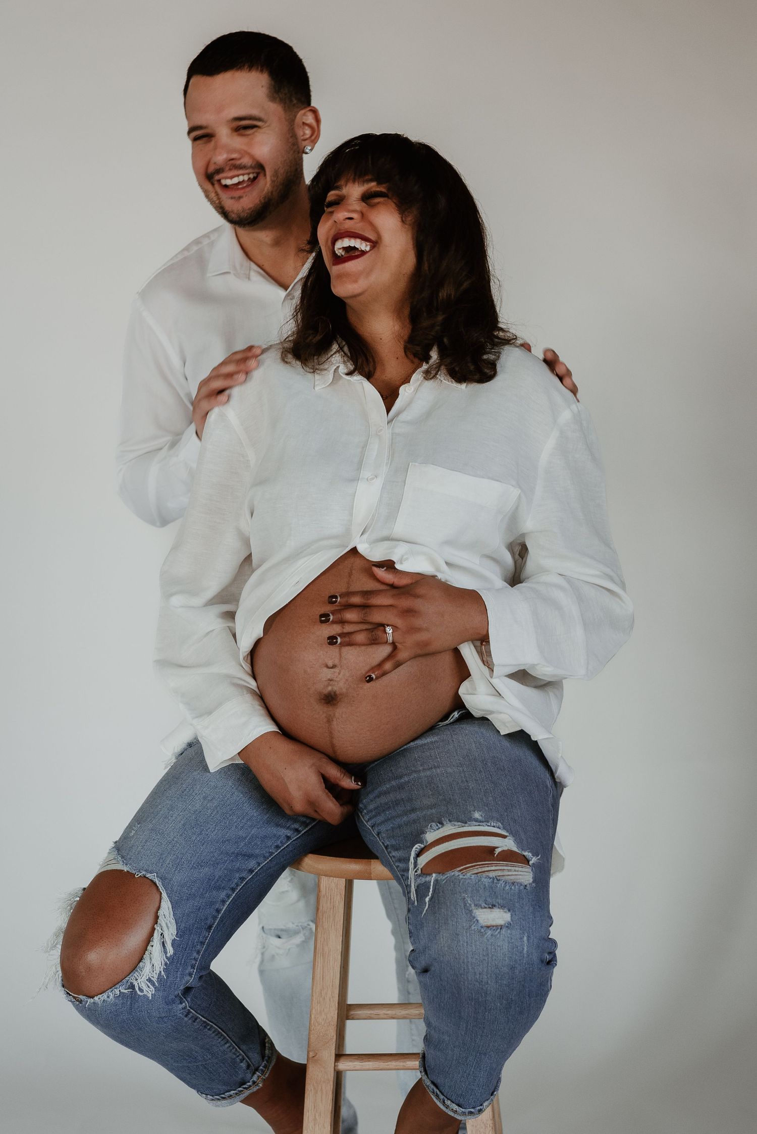 Joyful maternity photoshoot with couple laughing and embracing on wooden stool.