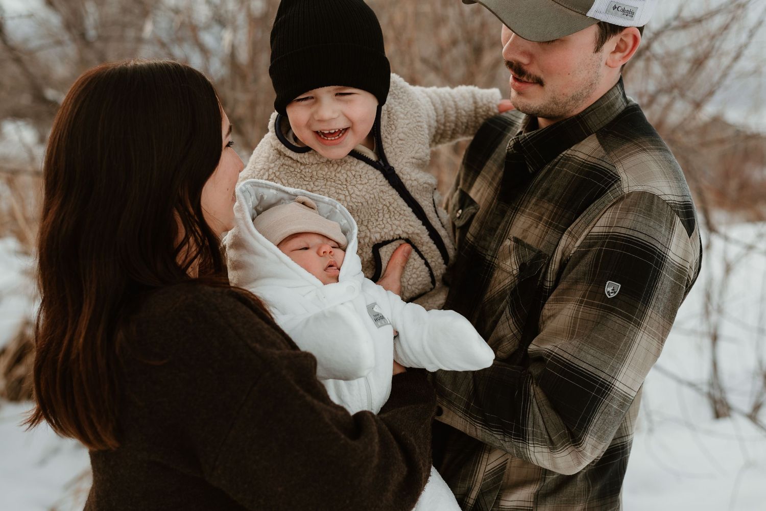 A family shares a tender winter moment outdoors while holding their newborn baby wrapped in white blankets.