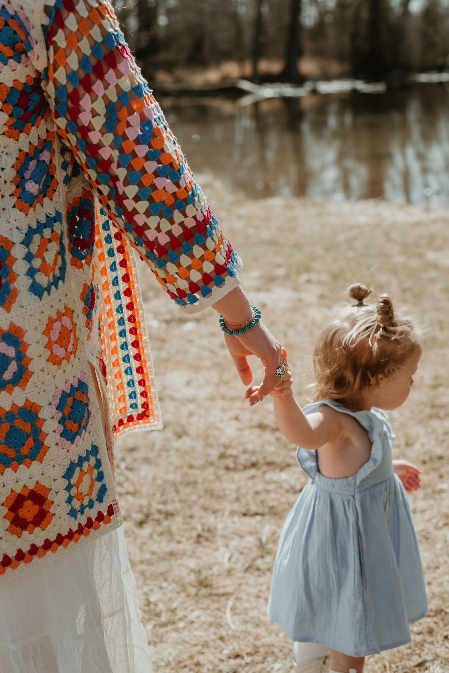 Someone in a blue dress reaches up toward a colorful crocheted cardigan in an outdoor setting.
