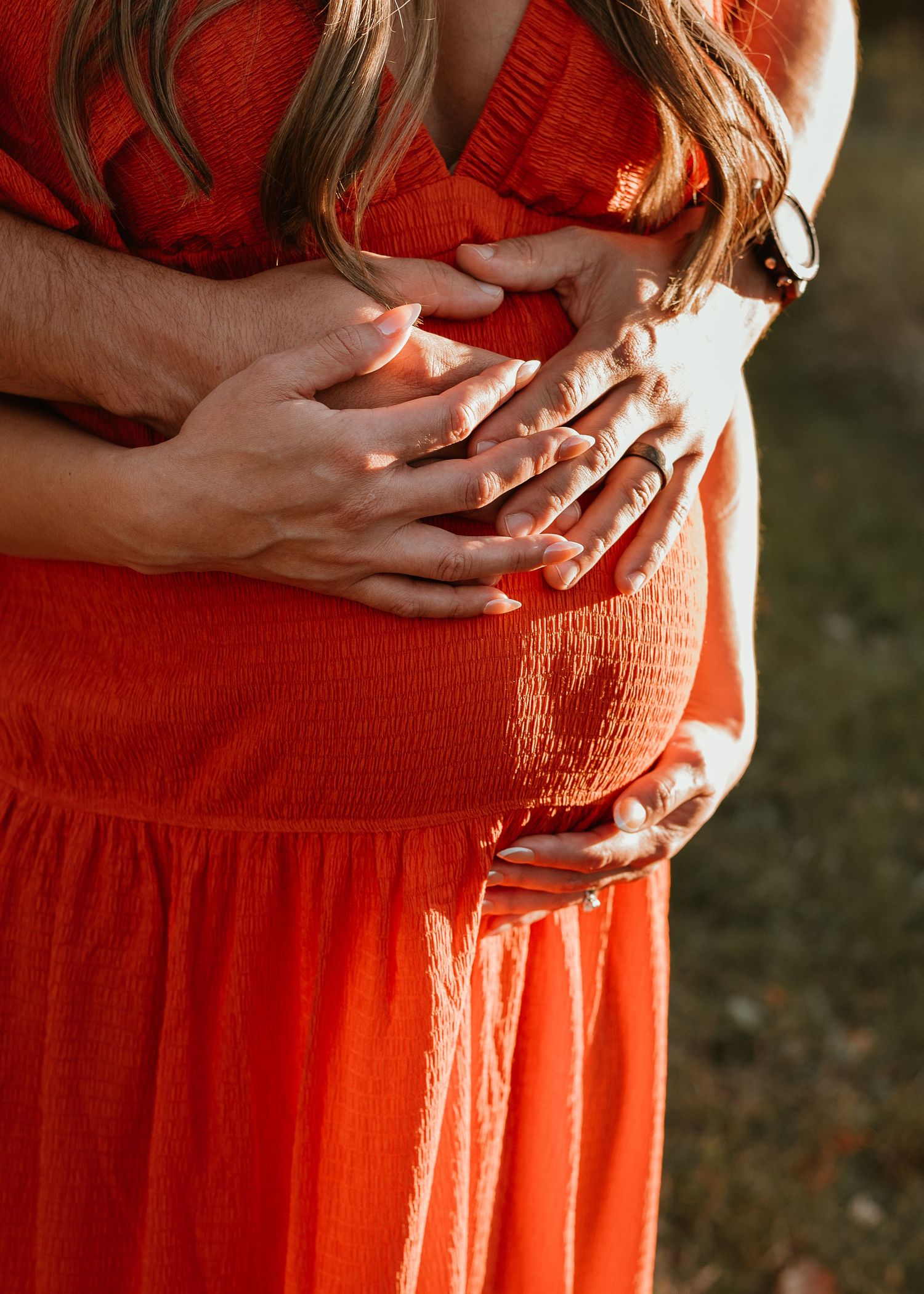 Hands tenderly rest on a pregnant belly draped in a vibrant red-orange flowing dress in warm sunlight.