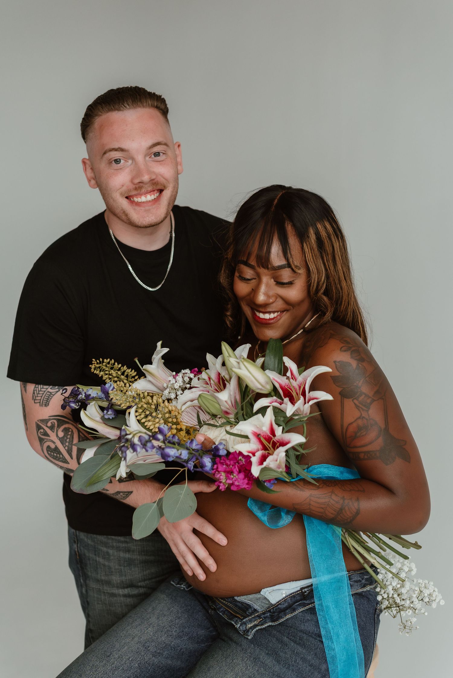 A couple laughs and embraces while holding a colorful floral bouquet with blue ribbon against a light gray backdrop.