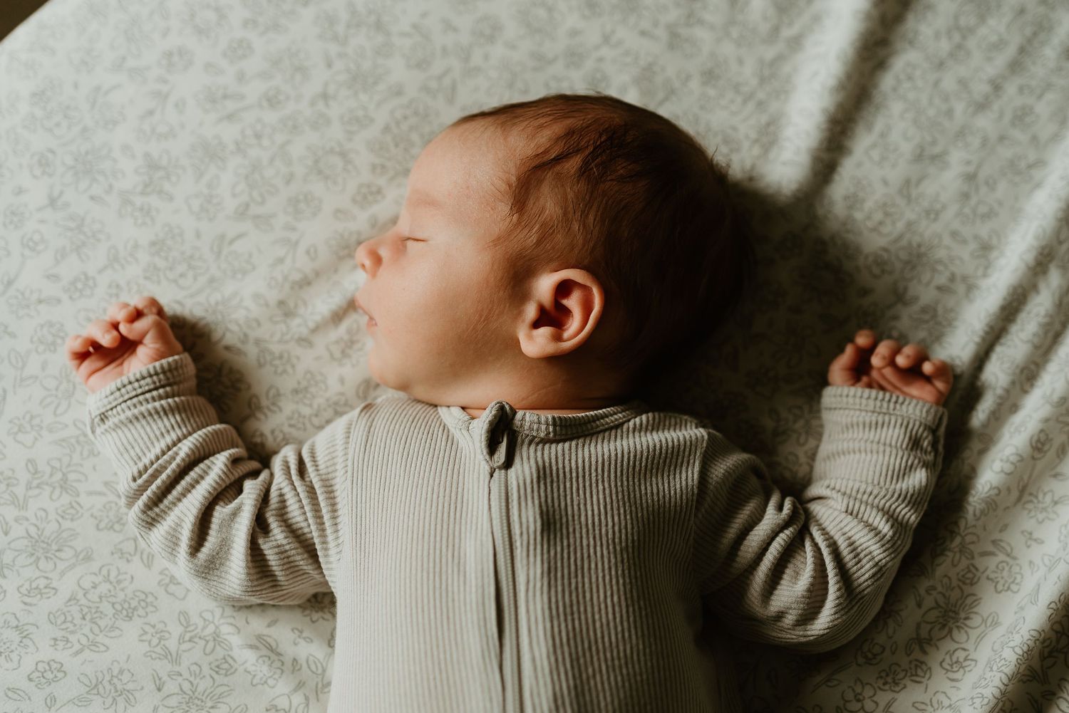 Baby in a gray onesie lies on a gray blanket with arms outstretched in a peaceful sleeping position.