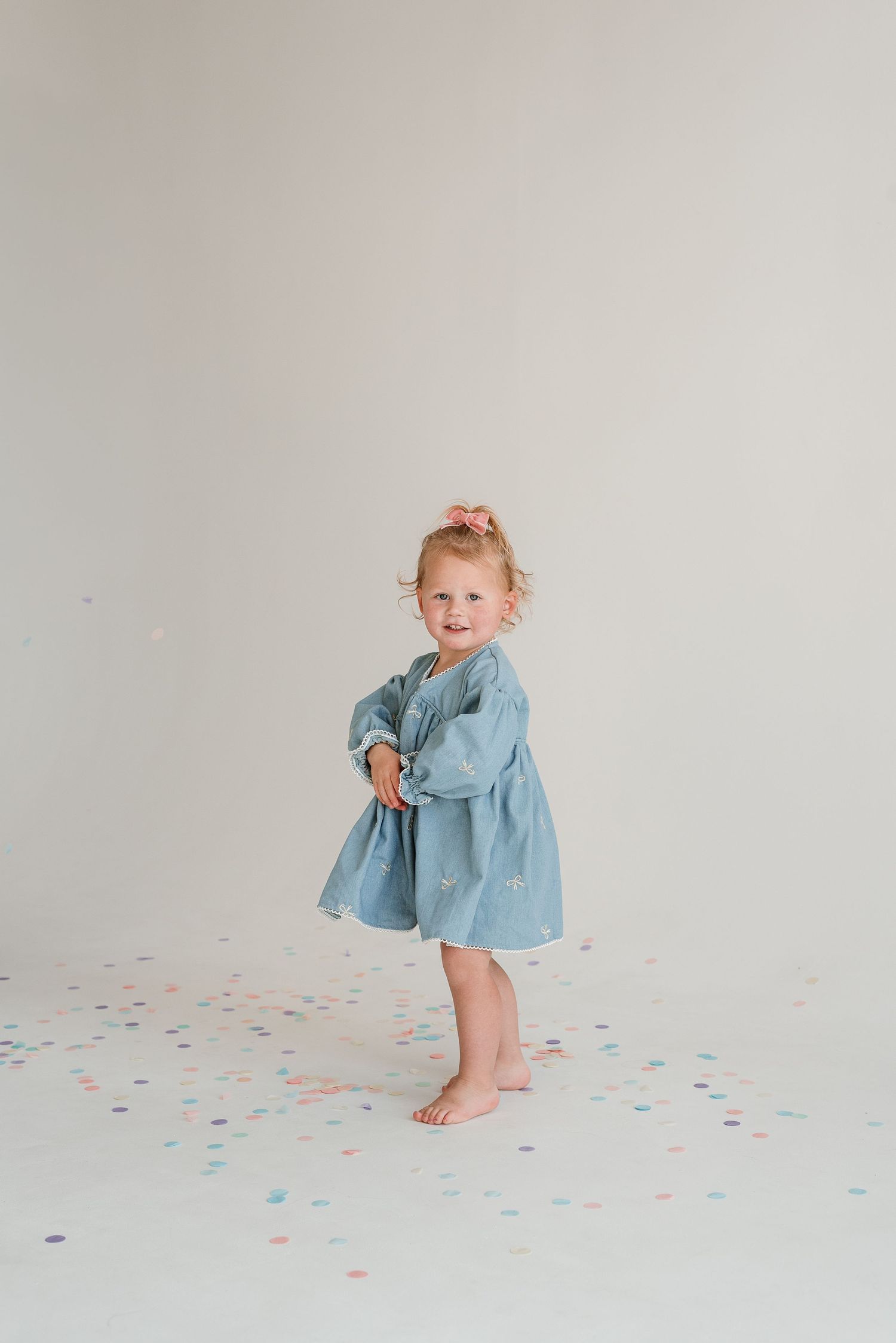Small child in light blue dress plays with confetti on white floor in minimalist photo studio setting.