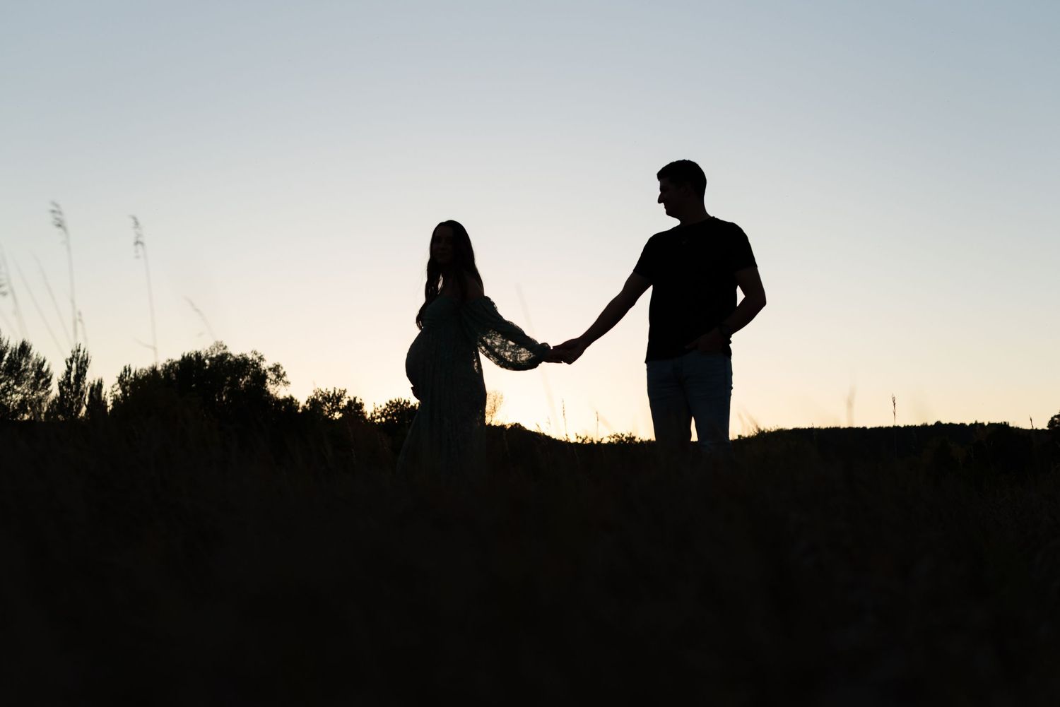 Silhouetted figures hold hands while walking along a hilltop ridge against a bright evening sky.