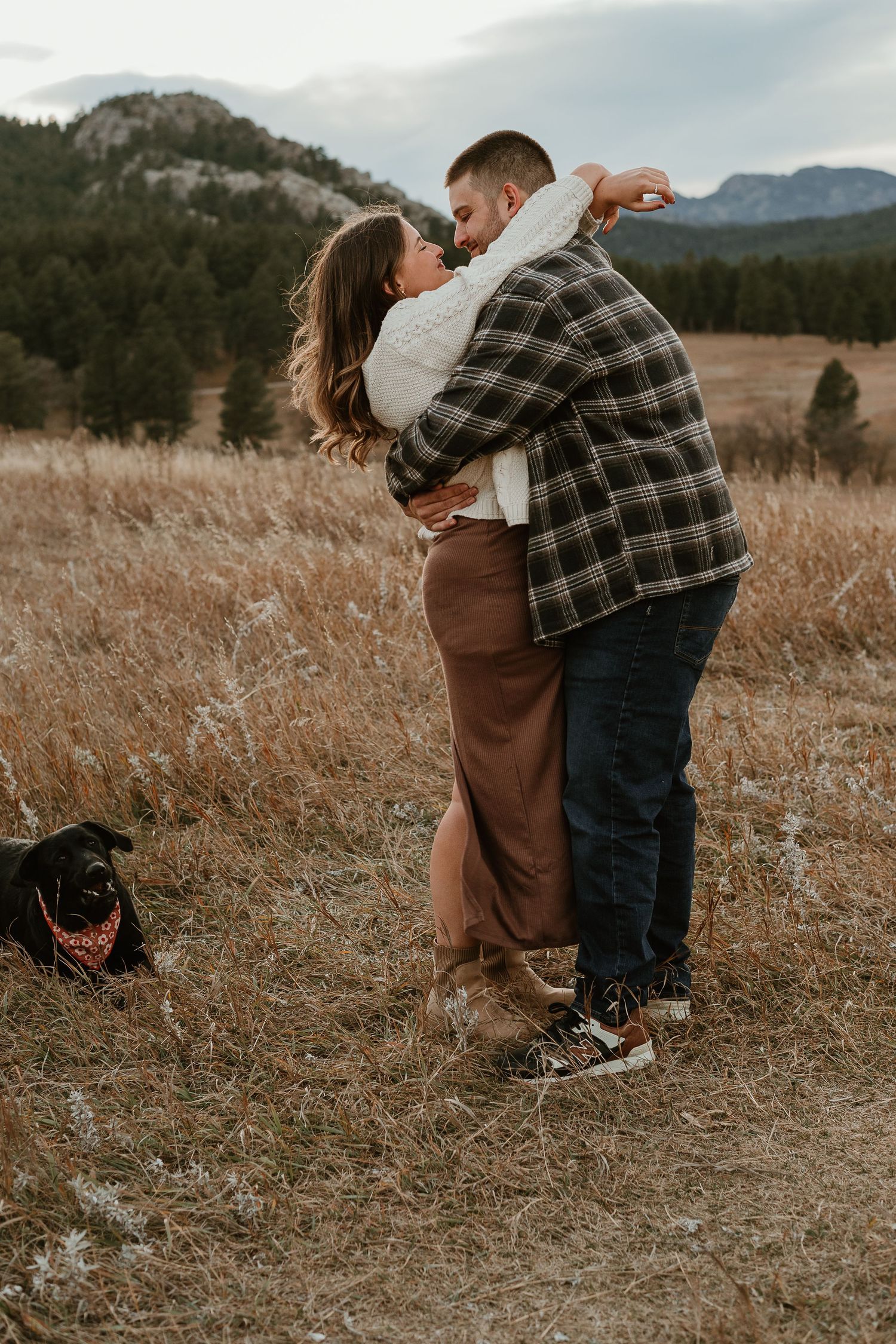 Romantic embrace in a mountain meadow with black dog watching nearby.