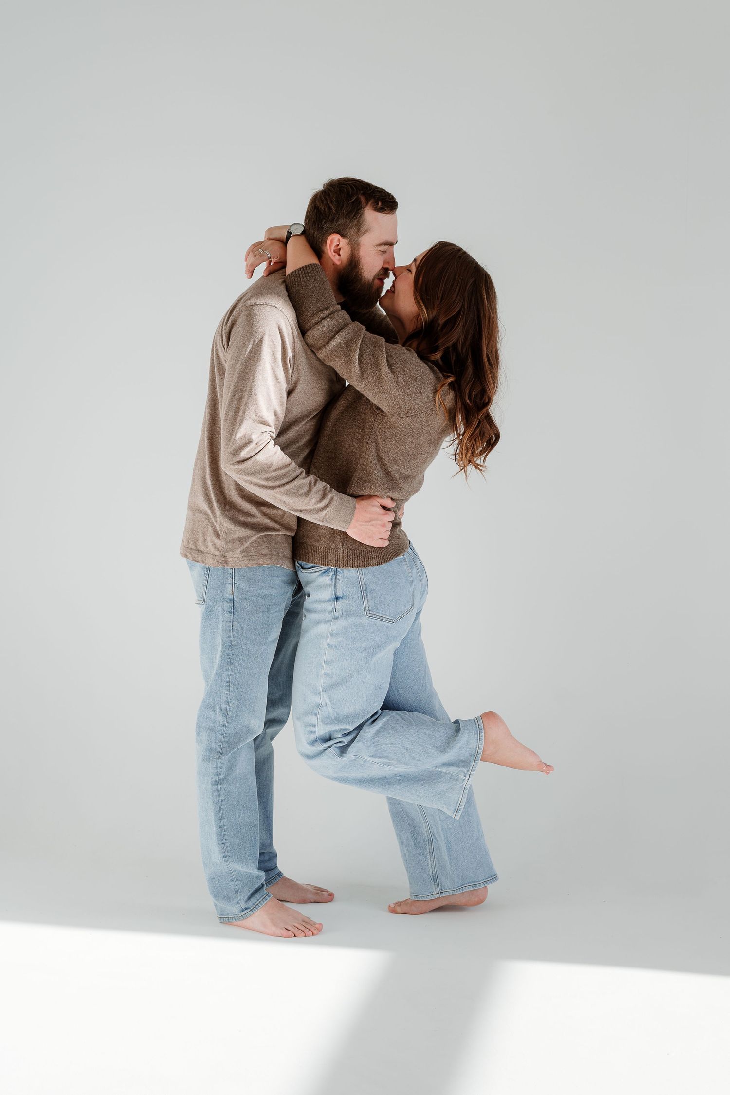 A couple in casual clothing sharing an intimate embrace and kiss against a plain white background.