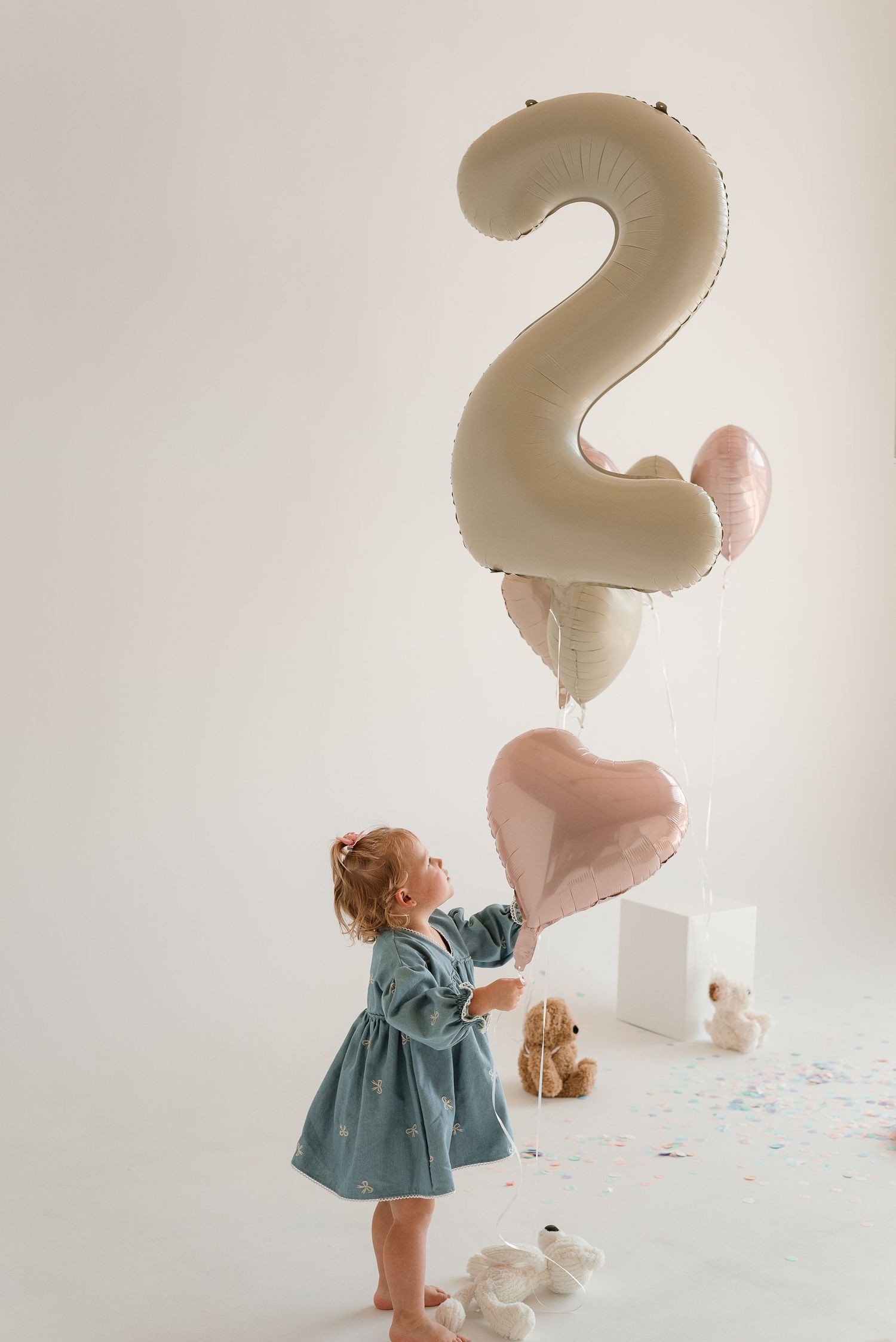 Second birthday photo with toddler in blue dress reaching for heart balloons and number 2 decoration.