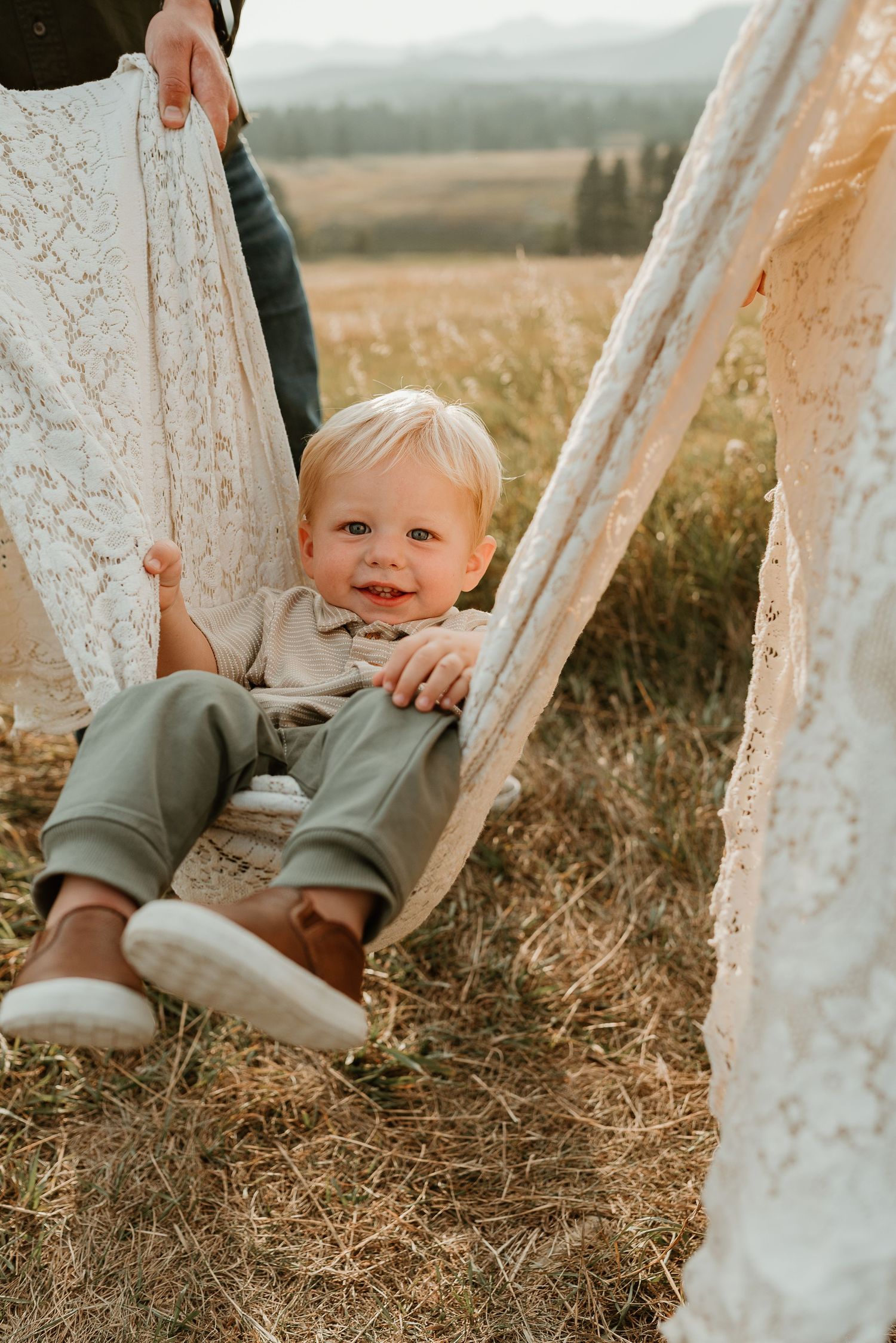 A child's feet dangle playfully between white lace curtains in an outdoor rustic setting.