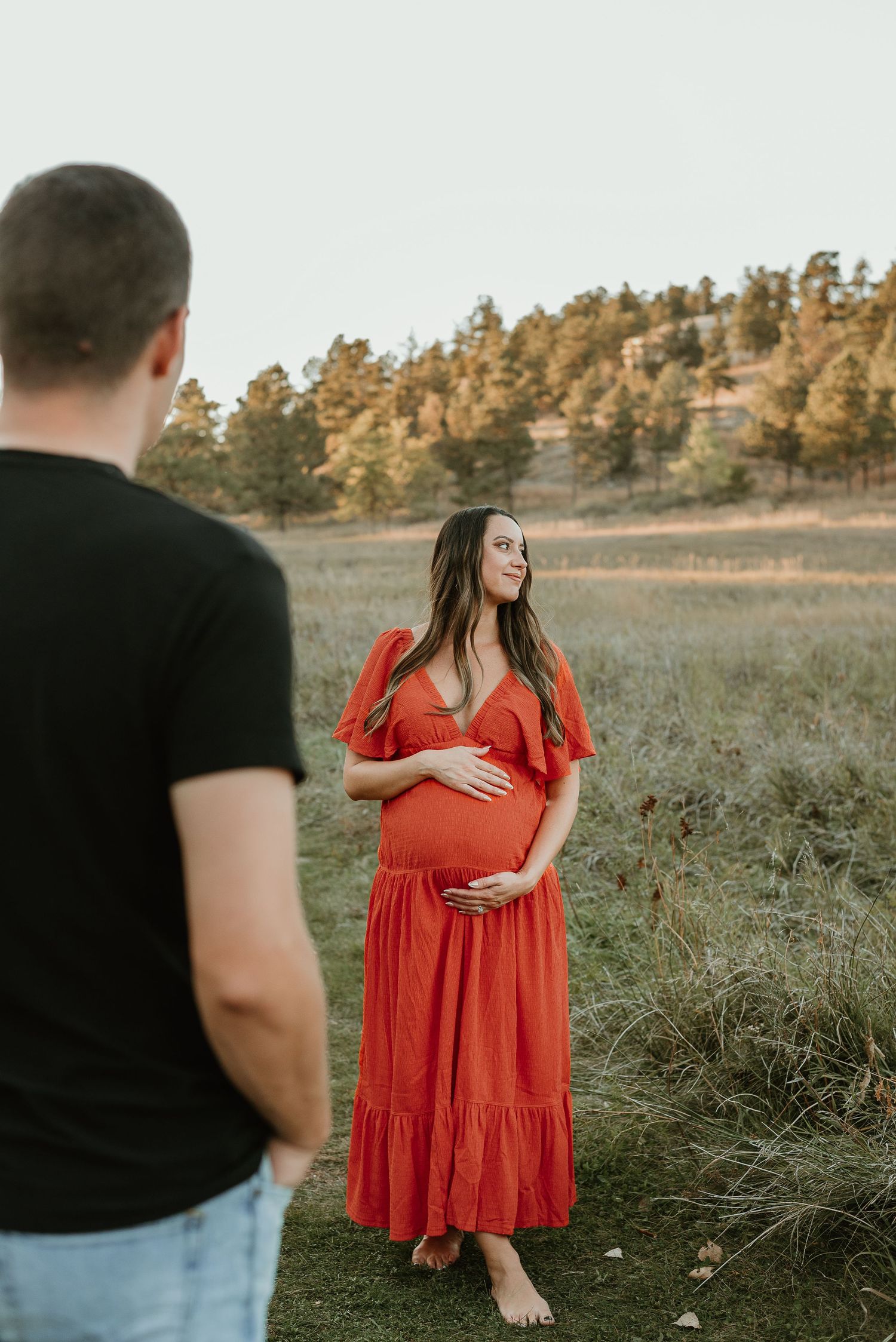 A maternity photoshoot in a field at sunset with a flowing red dress and mountain backdrop.