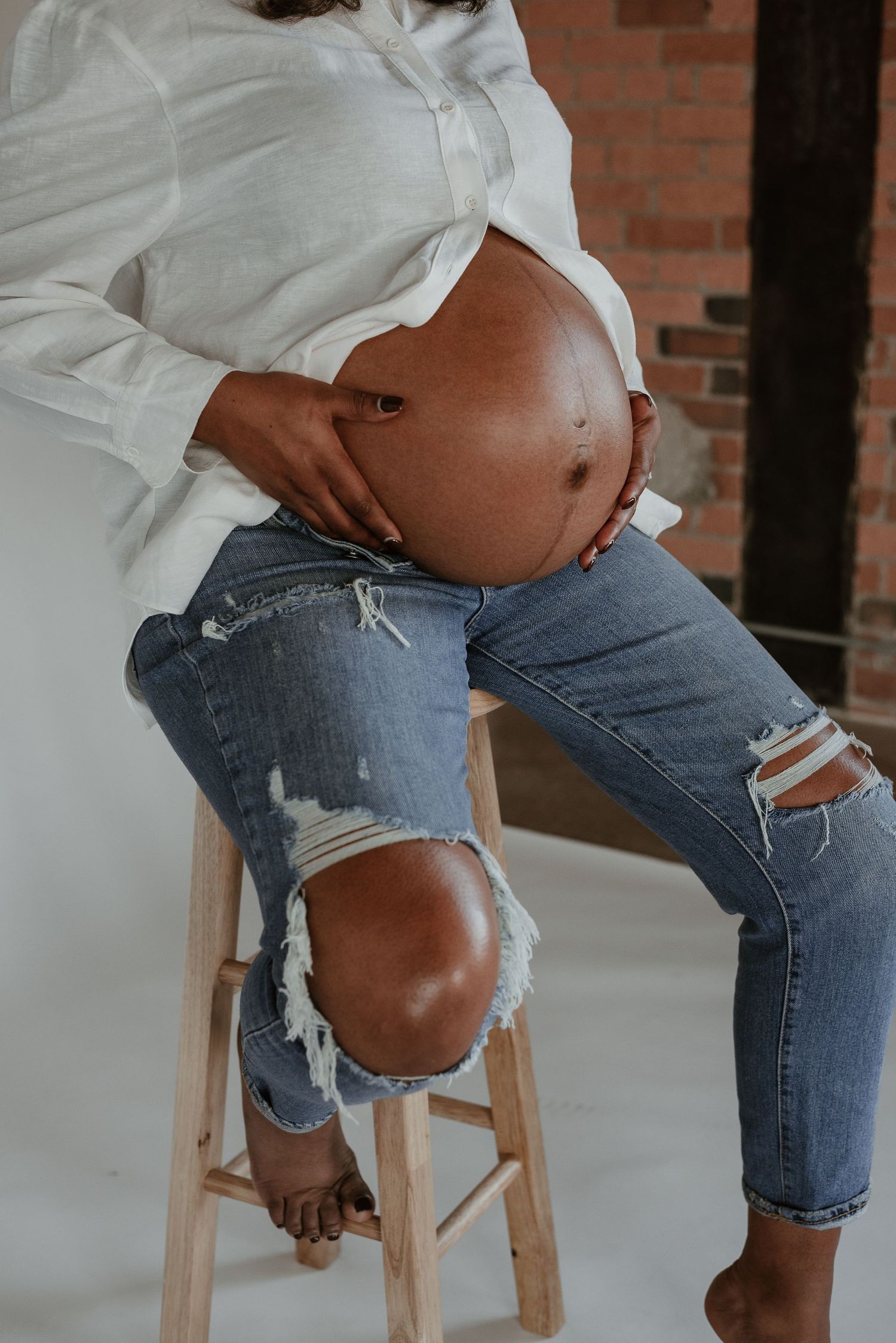 Close-up maternity photo showing pregnant belly with woman wearing distressed jeans on wooden stool.