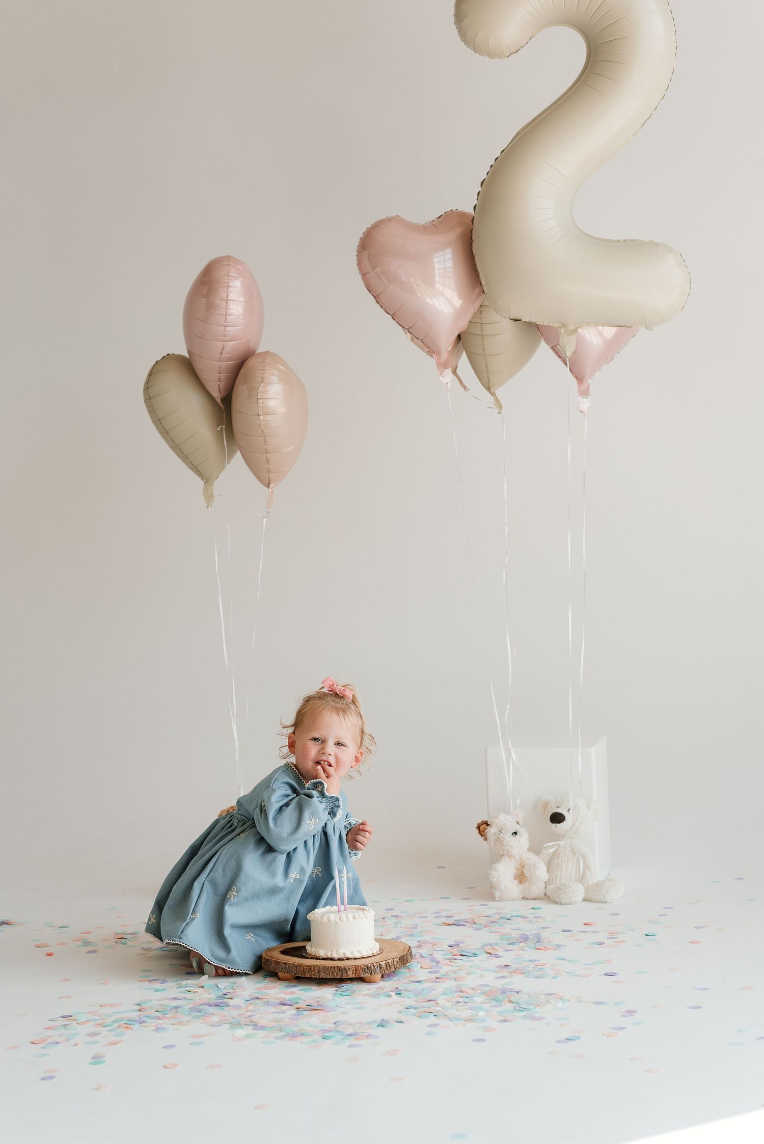 A baby in a blue dress sits on a white floor with heart-shaped balloons and a cake for their first birthday celebration.