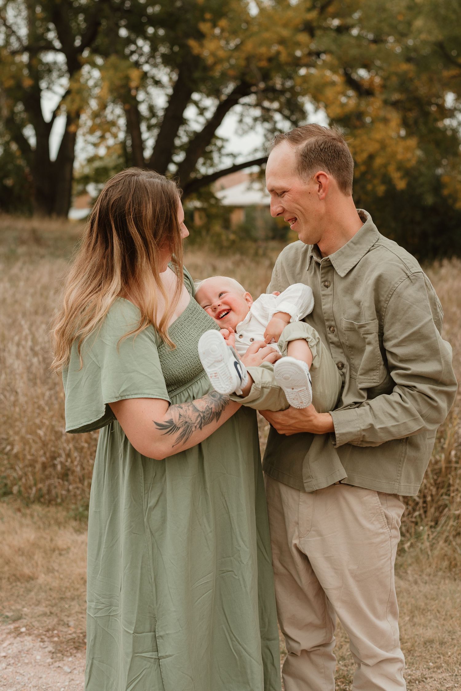 A couple in sage green and khaki outfits share joyful moments with their newborn baby in an autumn field setting.