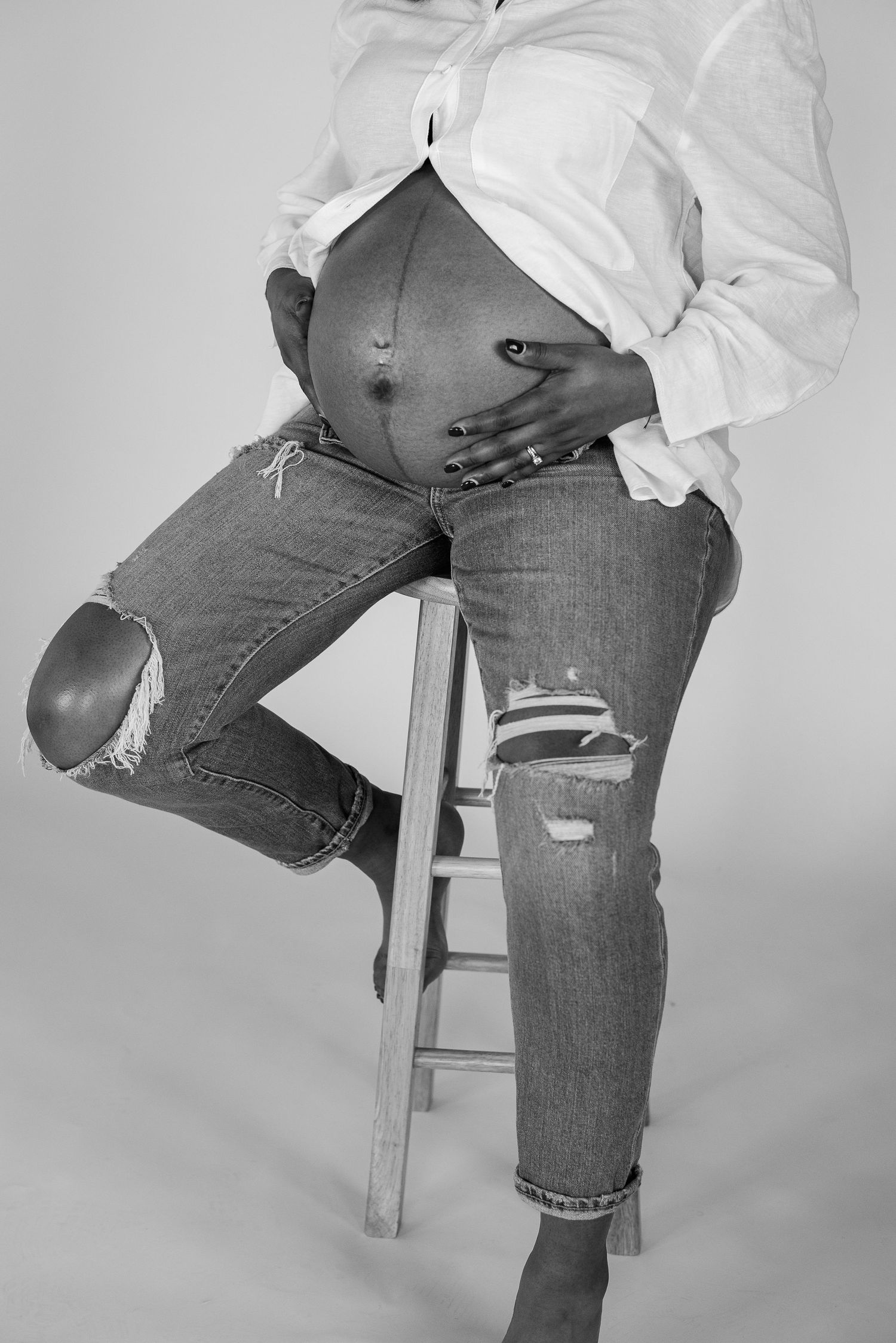 Black and white maternity portrait of pregnant woman seated on stool in casual attire.