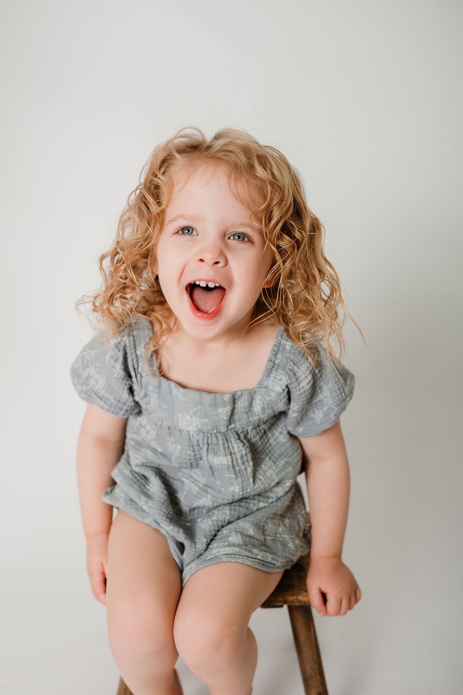 A child in a gray ruffled dress sits on a wooden chair with curly blonde hair and an expressive joyful smile.