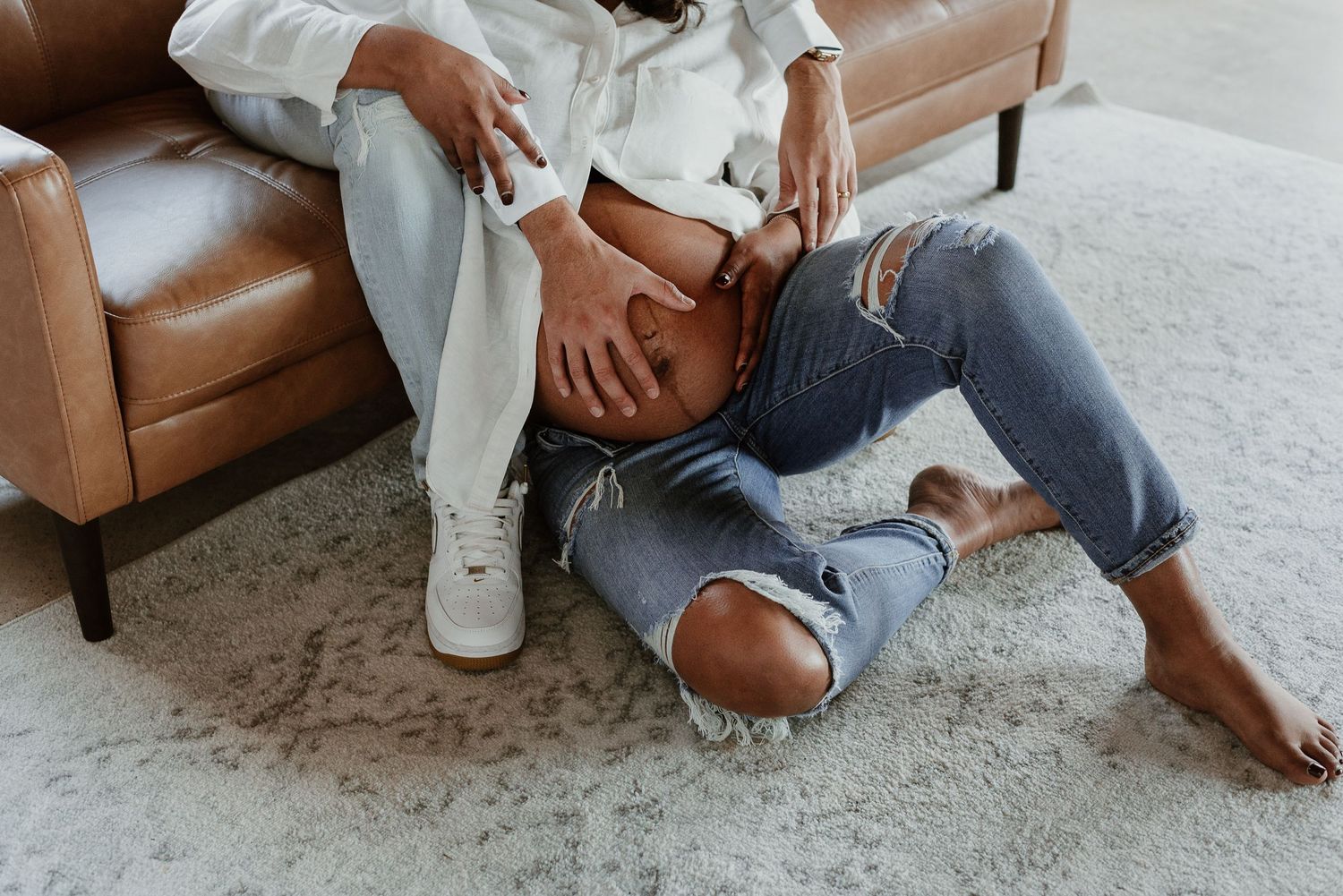 Casual lounging on carpet wearing distressed denim jeans, white sneakers and light colored top near brown leather sofa.