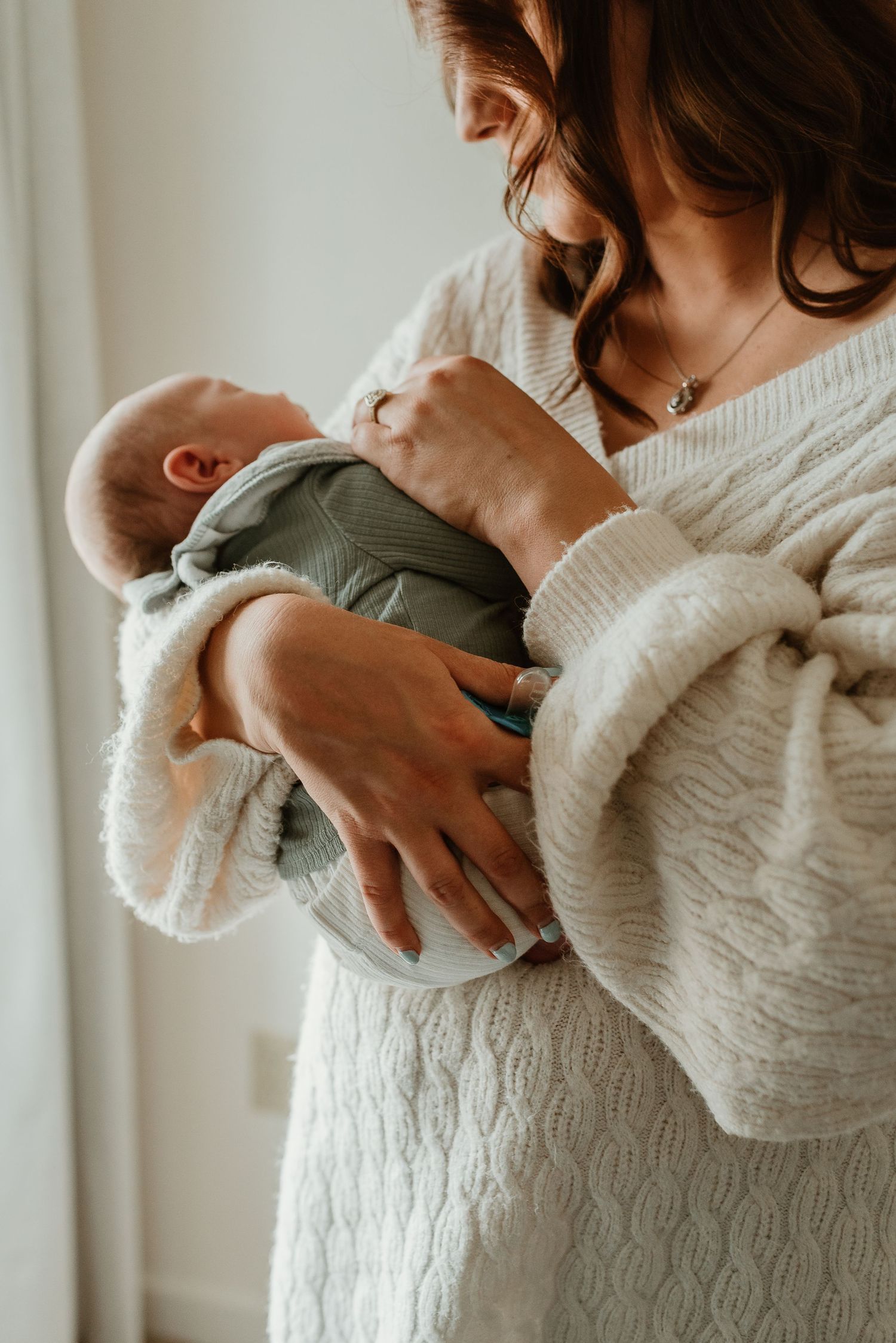 A tender moment showing a parent in a cream sweater cradling a baby wearing sage green against a light background.