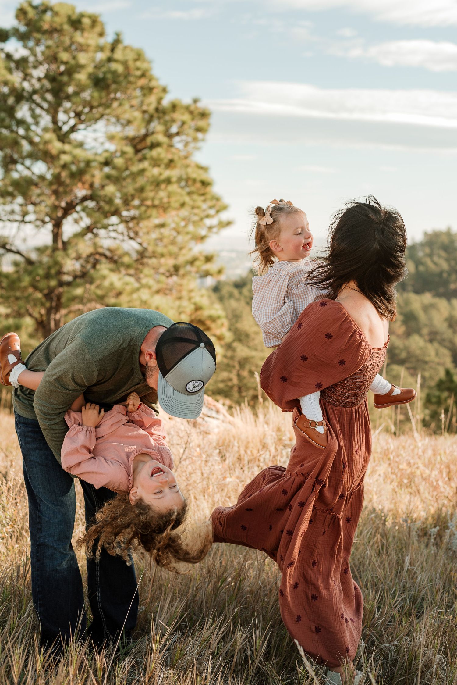 Couple in casual attire playfully dips and dances together in a sunlit field with tall grass and pine trees at sunset.