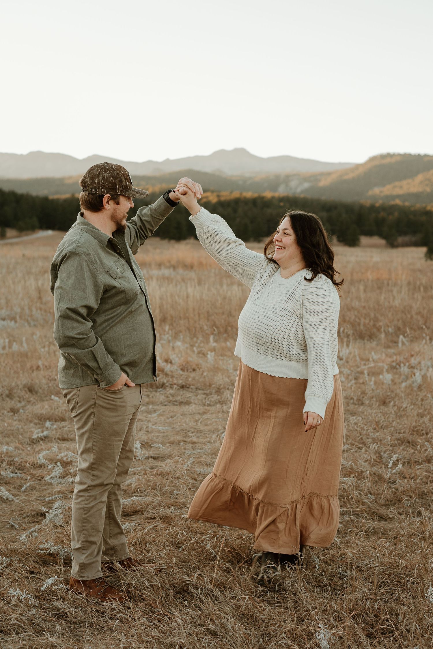 A couple shares playful moments in a golden field at sunset with mountains in the background.