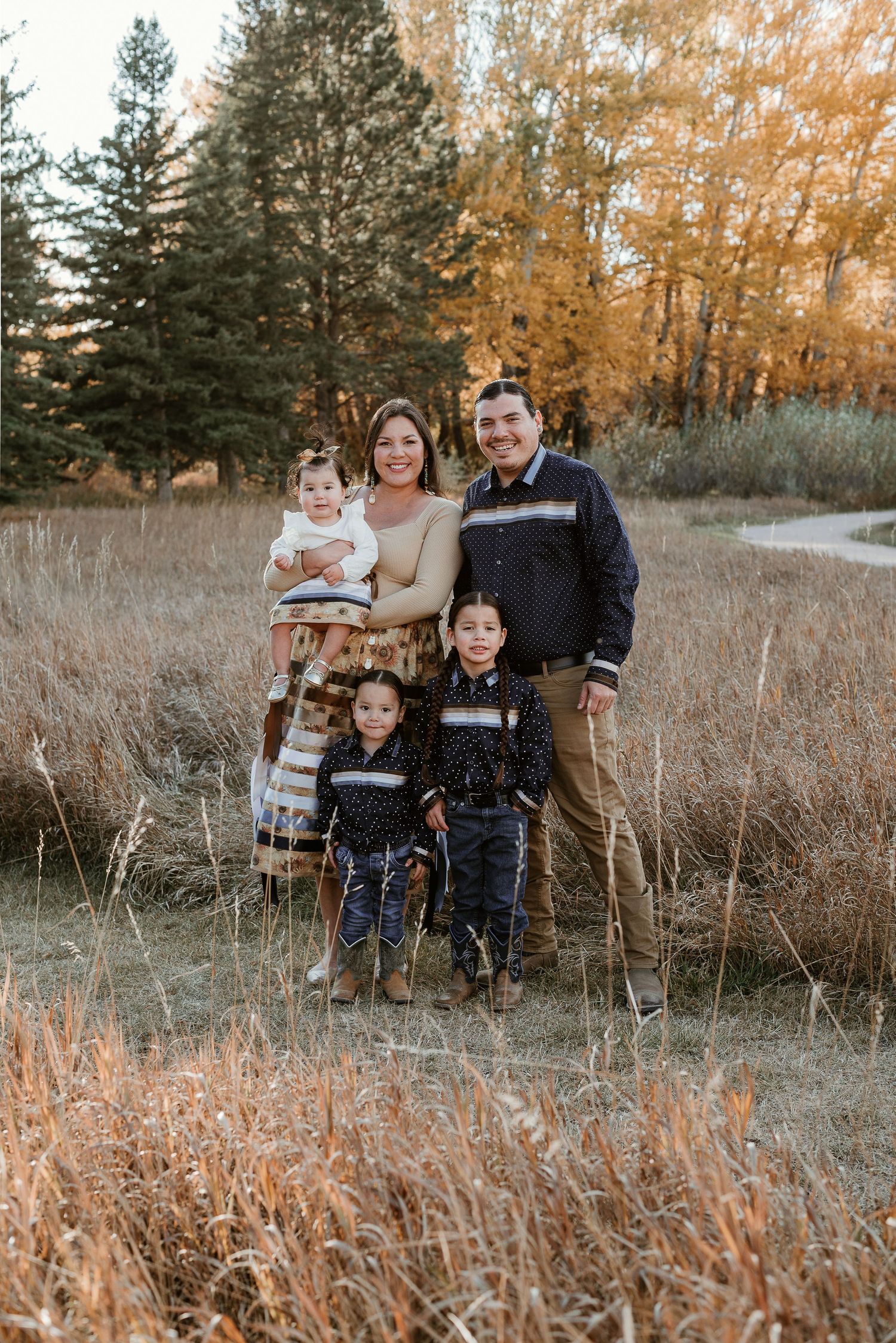 Family enjoying a playful moment together in a golden autumn field with evergreen trees at sunset.