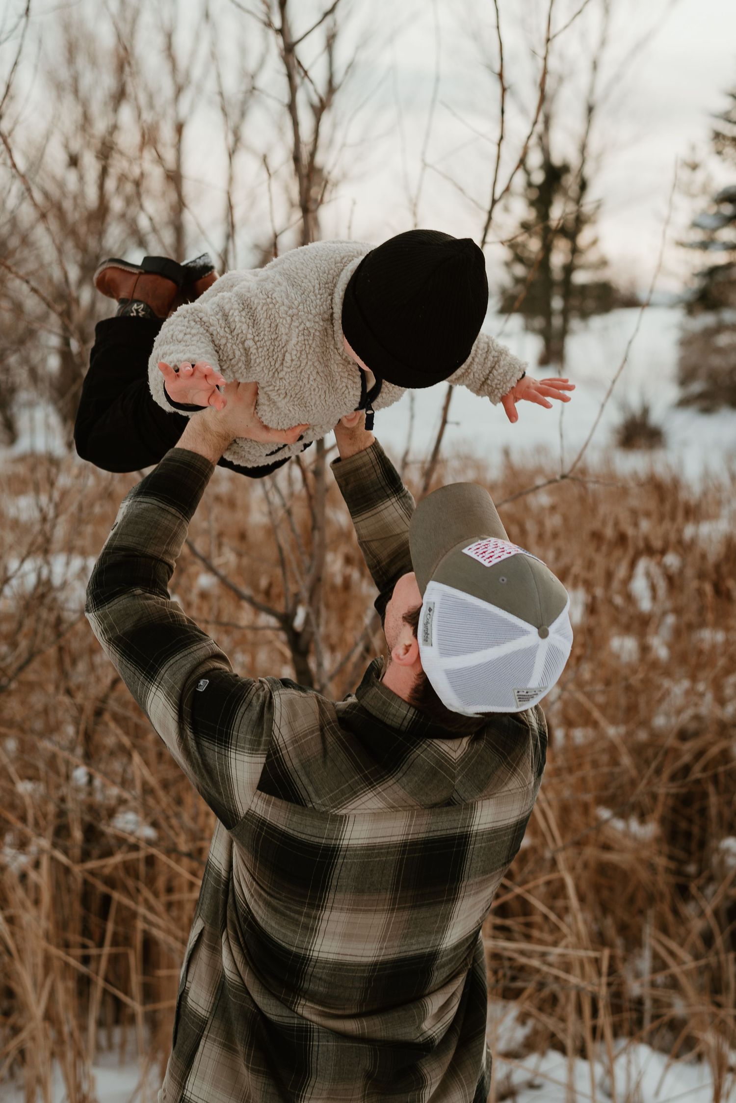 Parent in plaid shirt lifts child wearing winter clothing high in the air against snowy winter background.