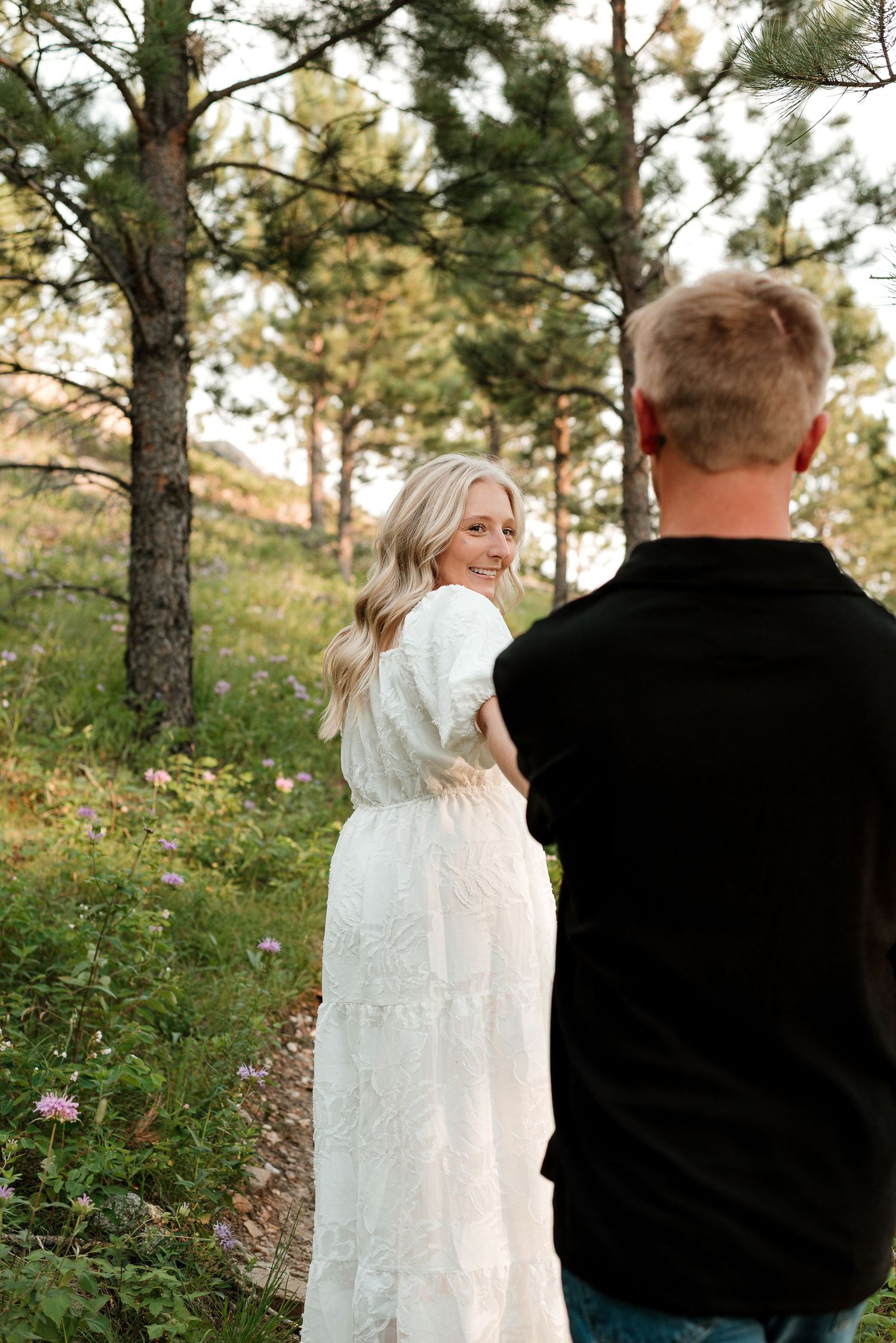 Couple in white dress and black shirt sharing intimate moment among pine trees.