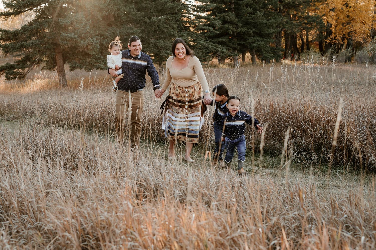 Family spending time together in tall grass field with evergreen trees during sunset.