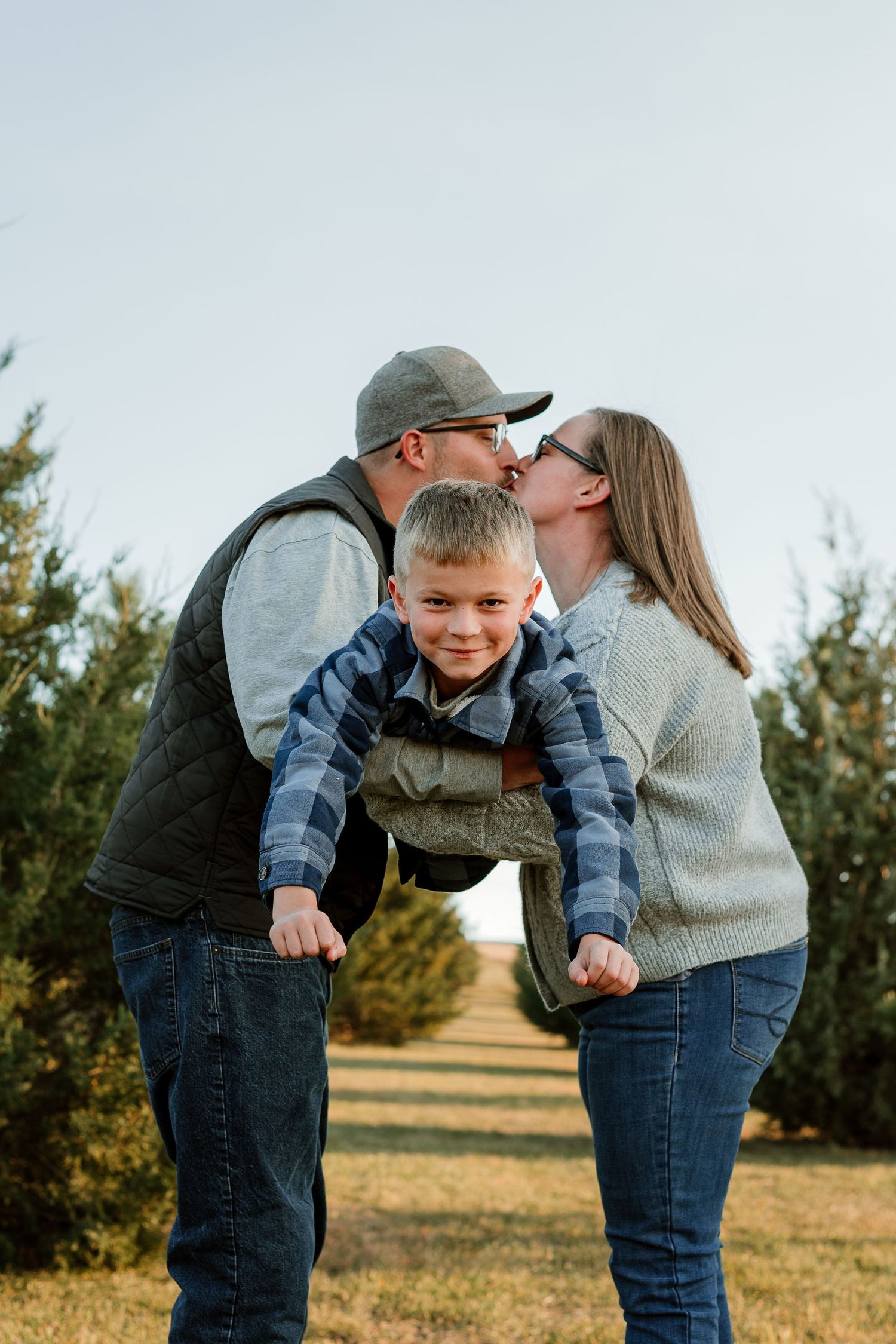 Parents share a kiss while playfully holding their young son between them in an outdoor setting at sunset.