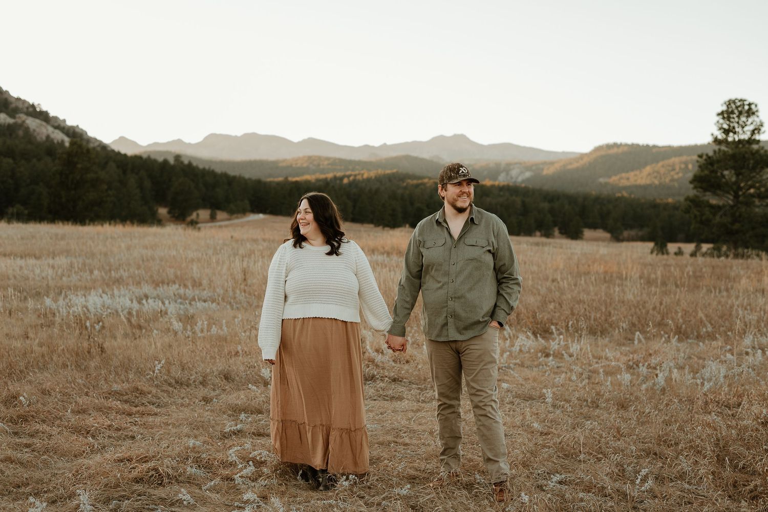 Couple walking hand in hand through a scenic mountain meadow at golden hour.