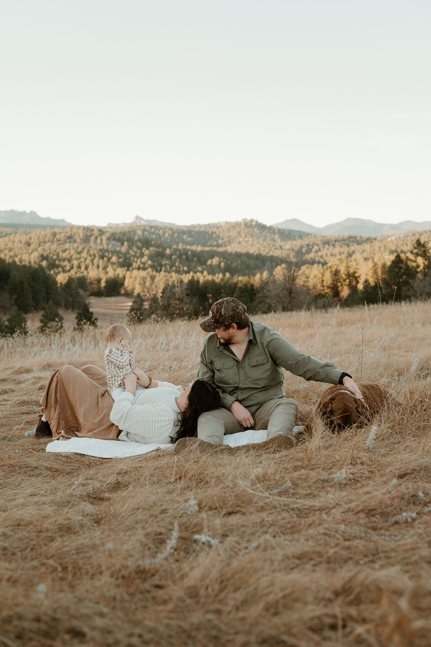 A couple lying together in a golden field at sunset with mountains in the background.