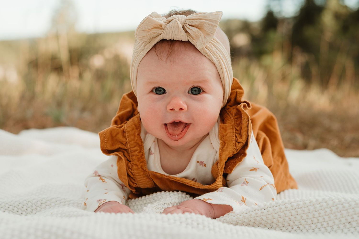 Baby lying on white blanket wearing brown clothing and smiling at camera.