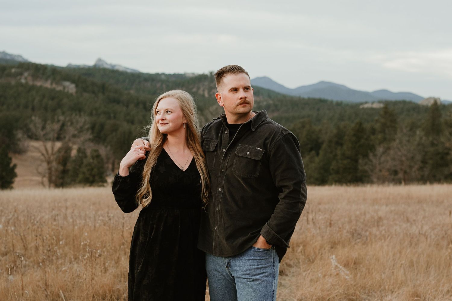 Two people stand together in a golden field with mountains and forest in the background on an overcast day.