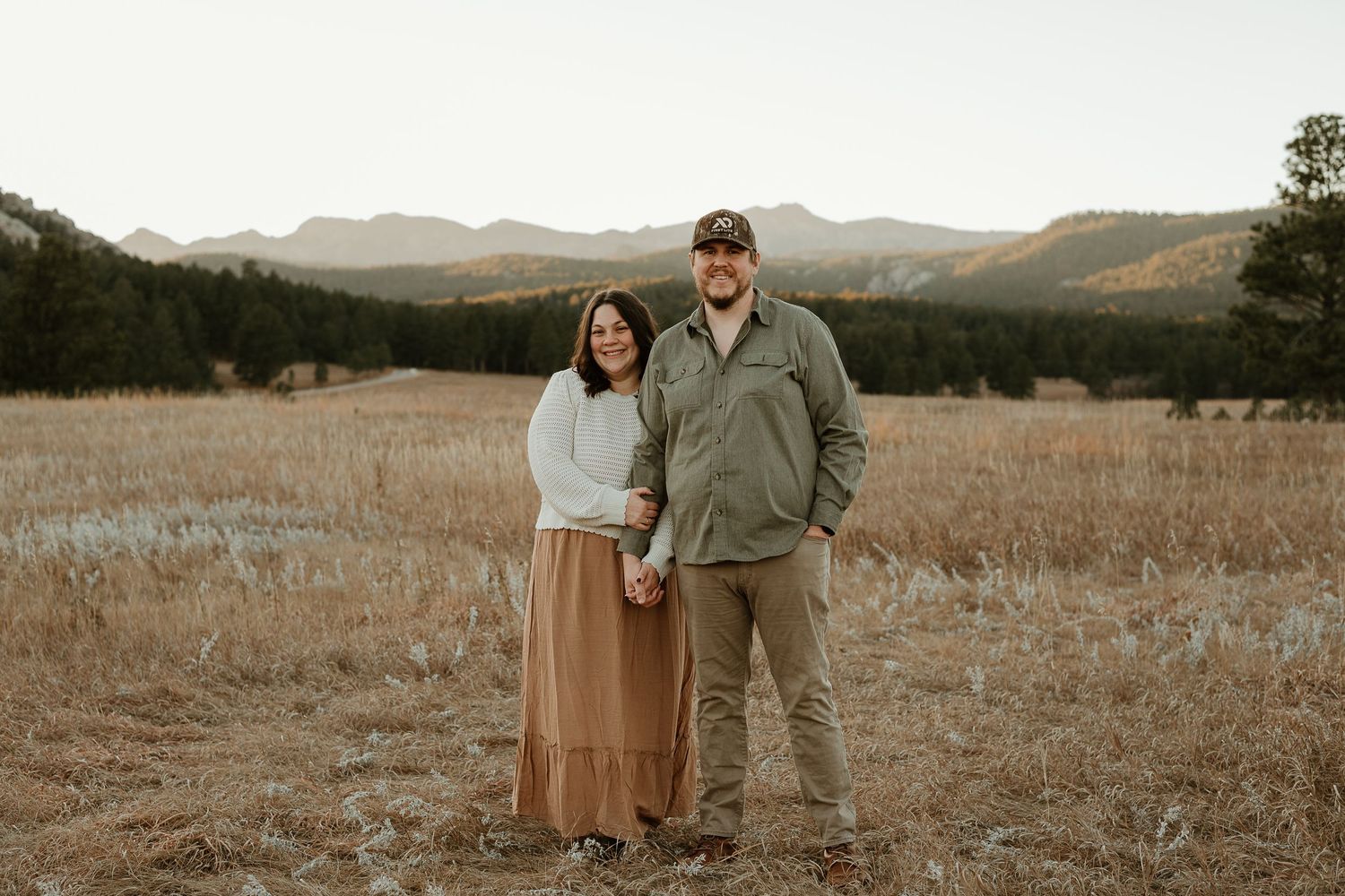 A couple embraces in a rustic field landscape with mountains and evergreen trees in the distance.