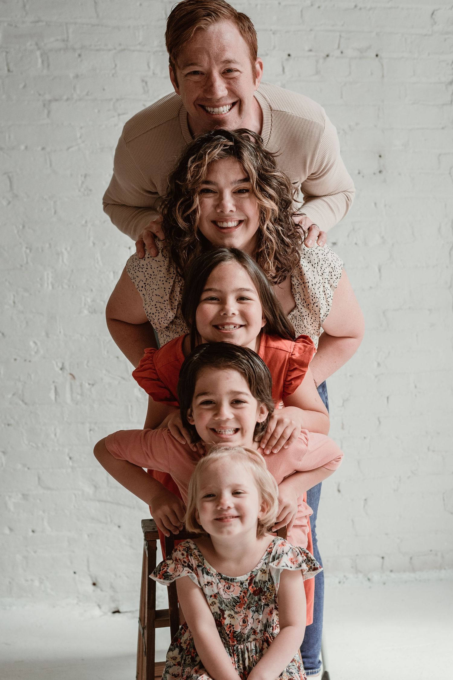 Family members stack together for a fun portrait against white brick wall.