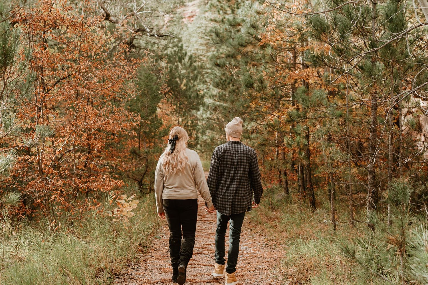 Two people walk hand in hand down a forest path surrounded by autumn foliage and evergreen trees.