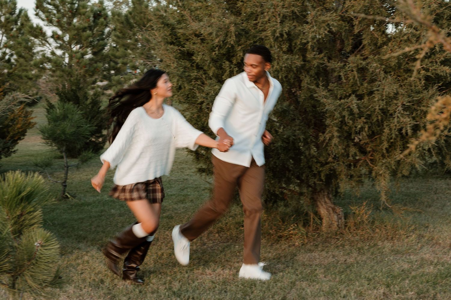 Couple playing and running together on grass near pine trees at sunset.