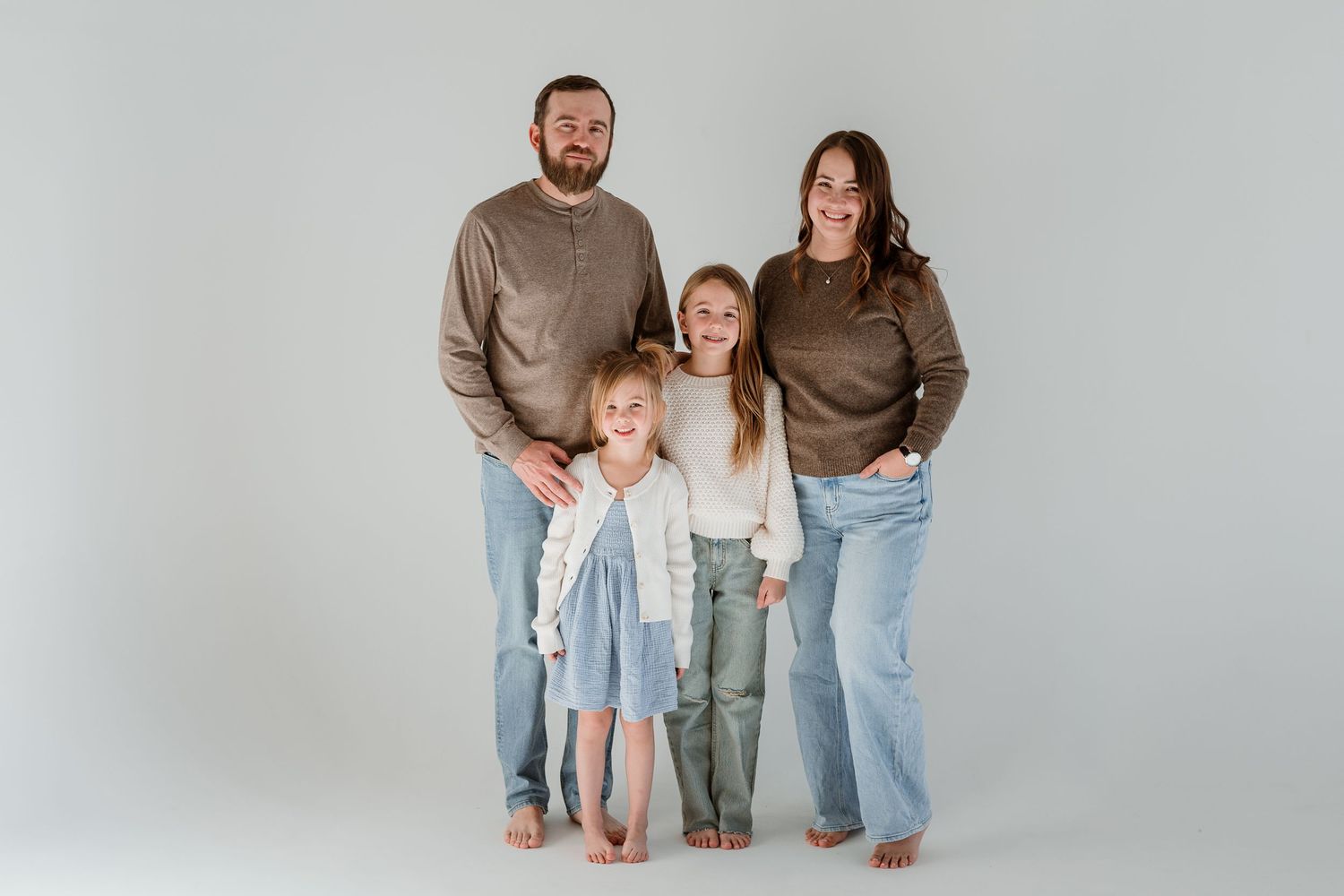Family wearing coordinated casual outfits in brown and blue tones poses together against white studio backdrop.
