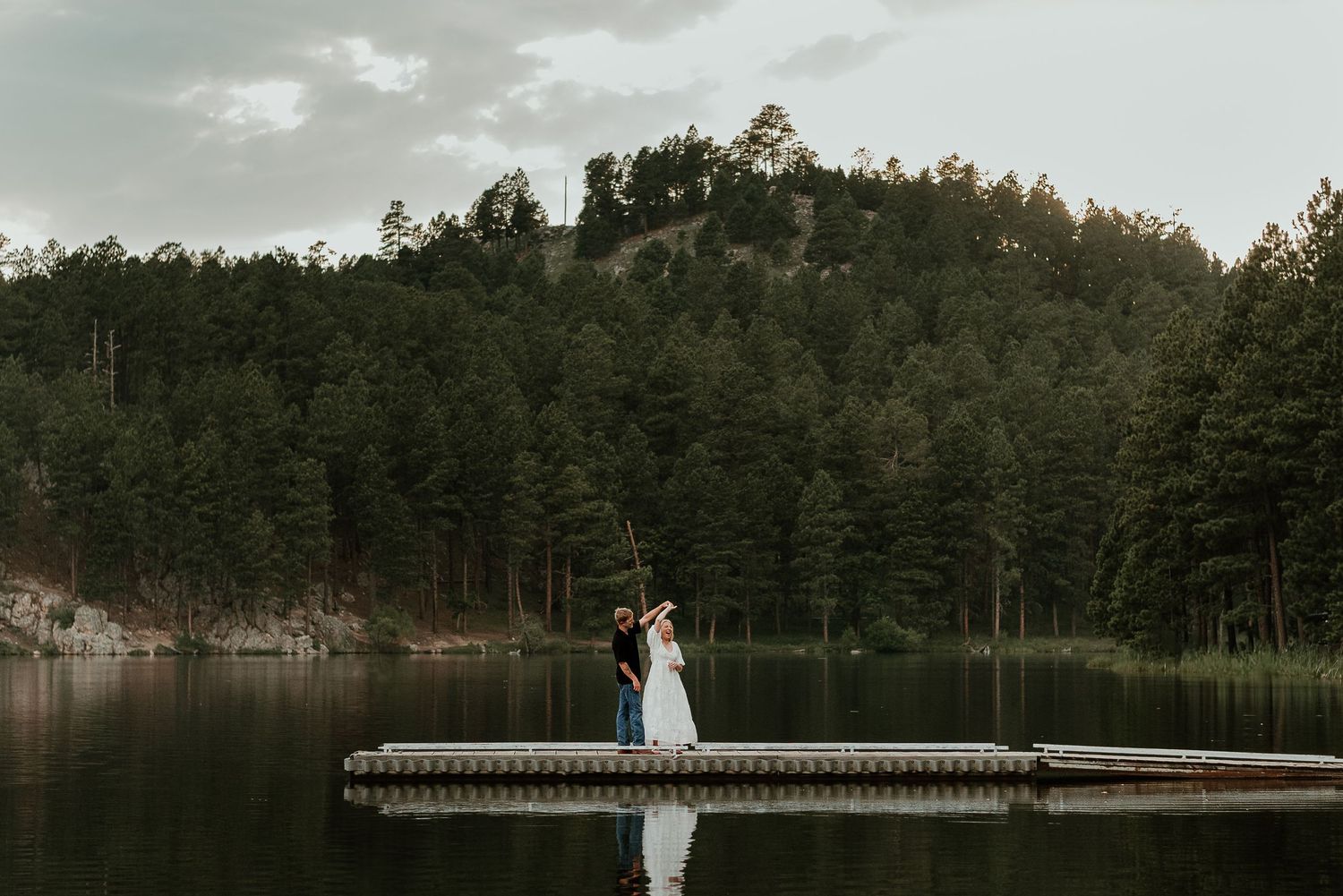 Panoramic mountain lake landscape with evergreen forest and wooden pier at sunset.