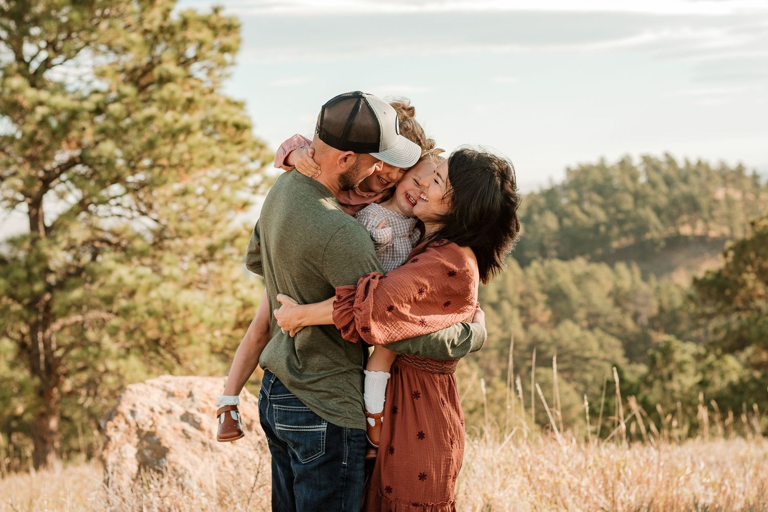 A couple embraces in a sun-drenched meadow with golden grass and evergreen trees in the background.