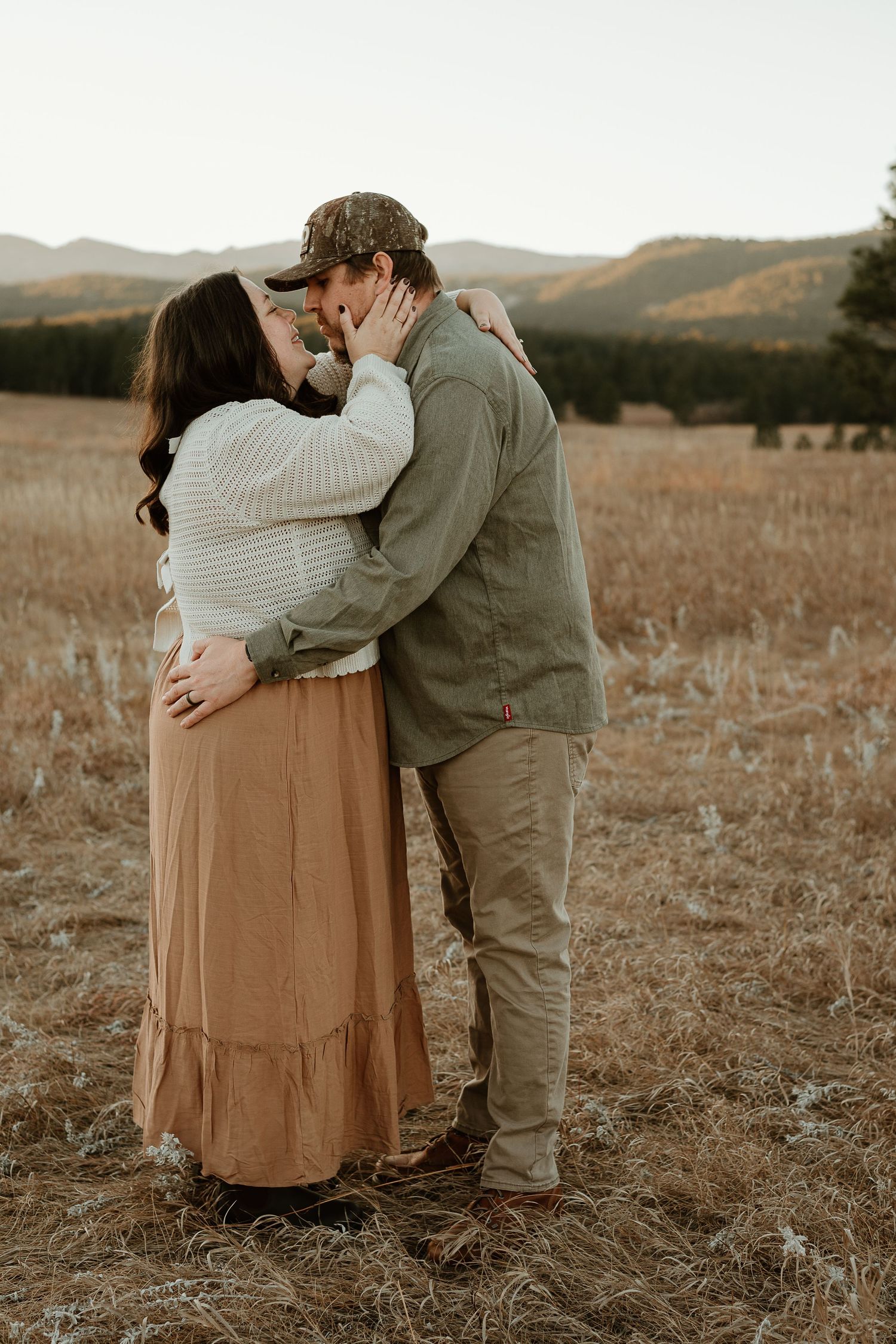 A romantic couple embraces while standing in a rustic field with mountains visible in the distance.