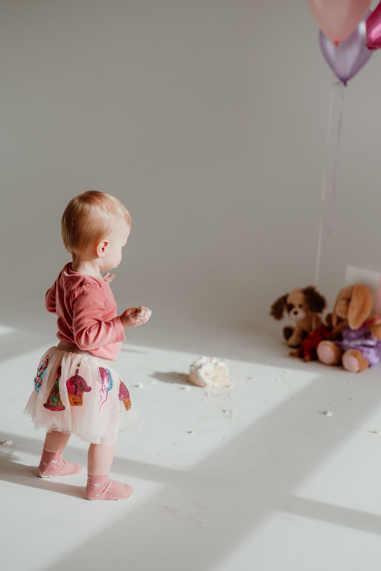 Toddler in pink top and tulle skirt stands near birthday decorations and stuffed animals.